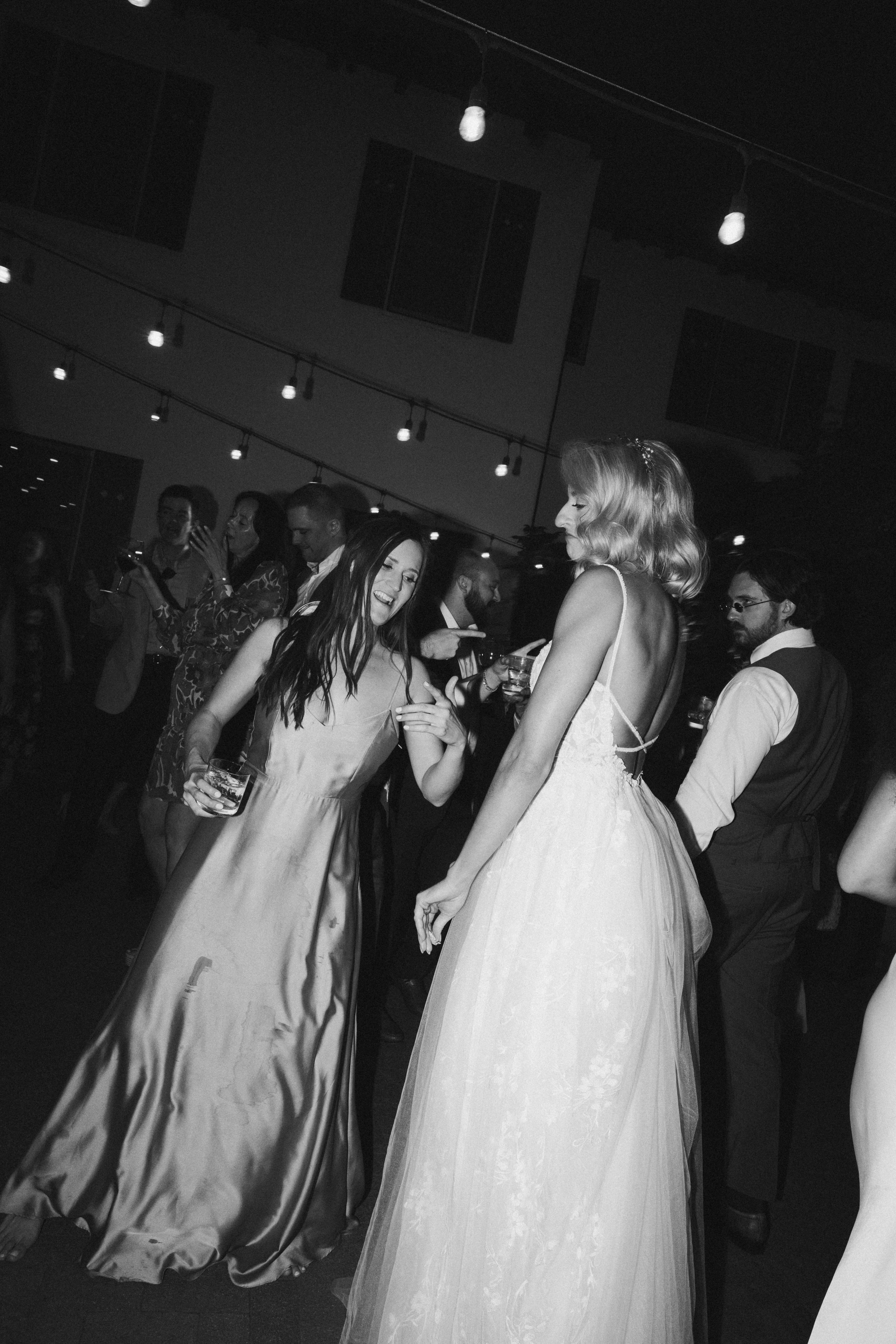 Two people in elegant dresses are dancing at an indoor event with string lights overhead. Others are visible in the background. Black and white image at quail ranch