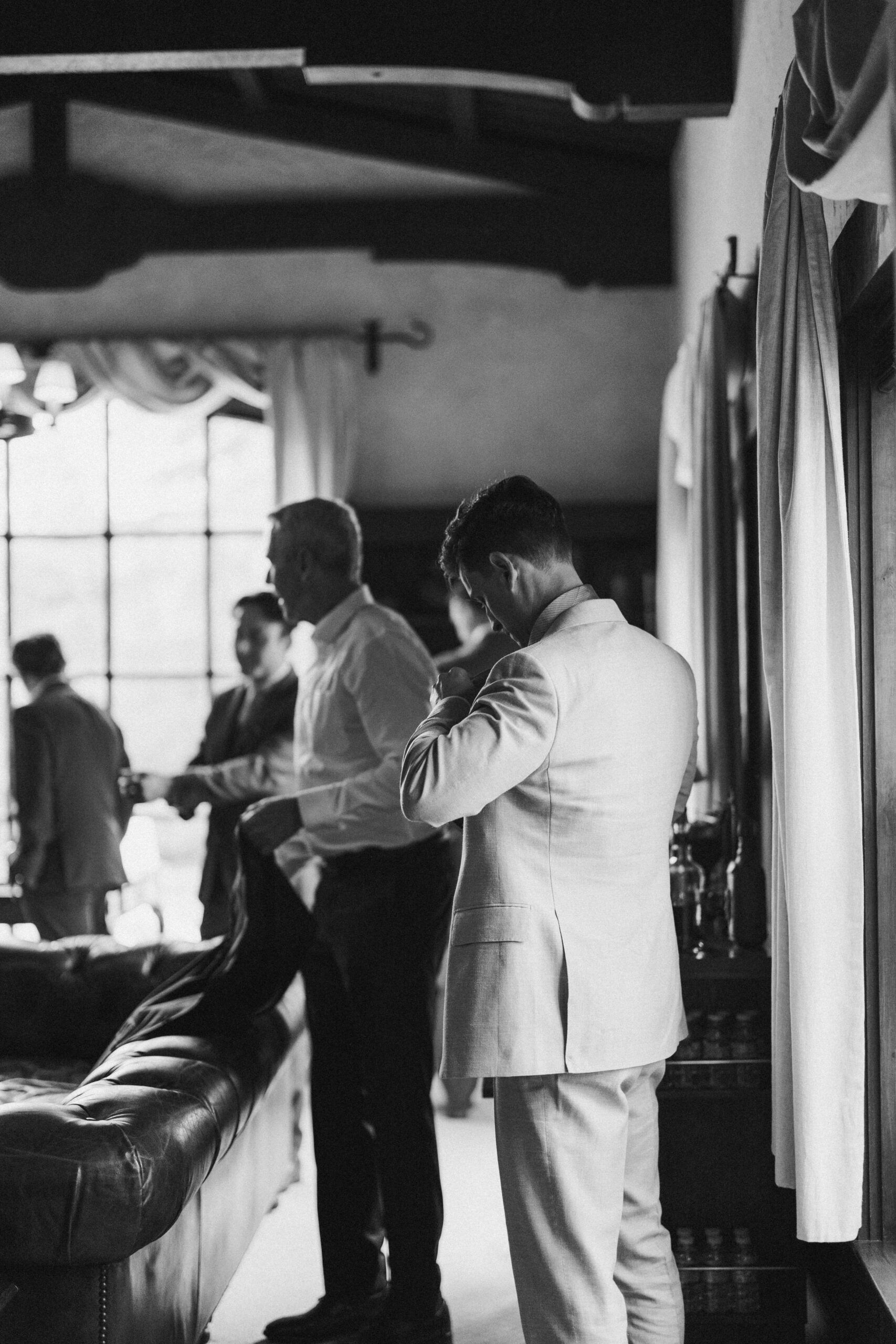 Black and white image of several men in formal attire in a room with large windows. One man adjusts his tie, while others engage in conversation.