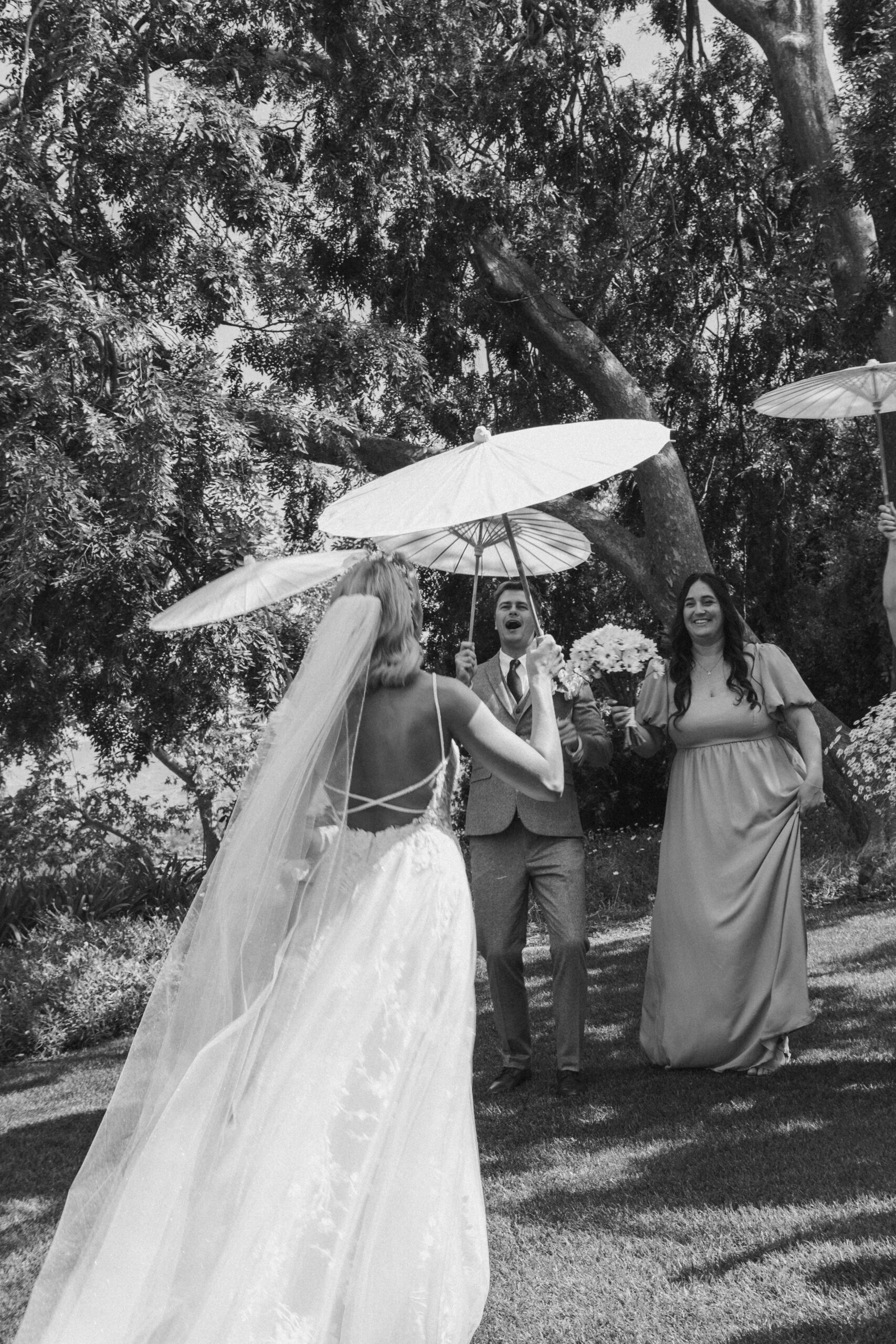 A bride holding an umbrella walks towards a man and a woman standing under umbrellas in a garden setting. The woman holds a bouquet.