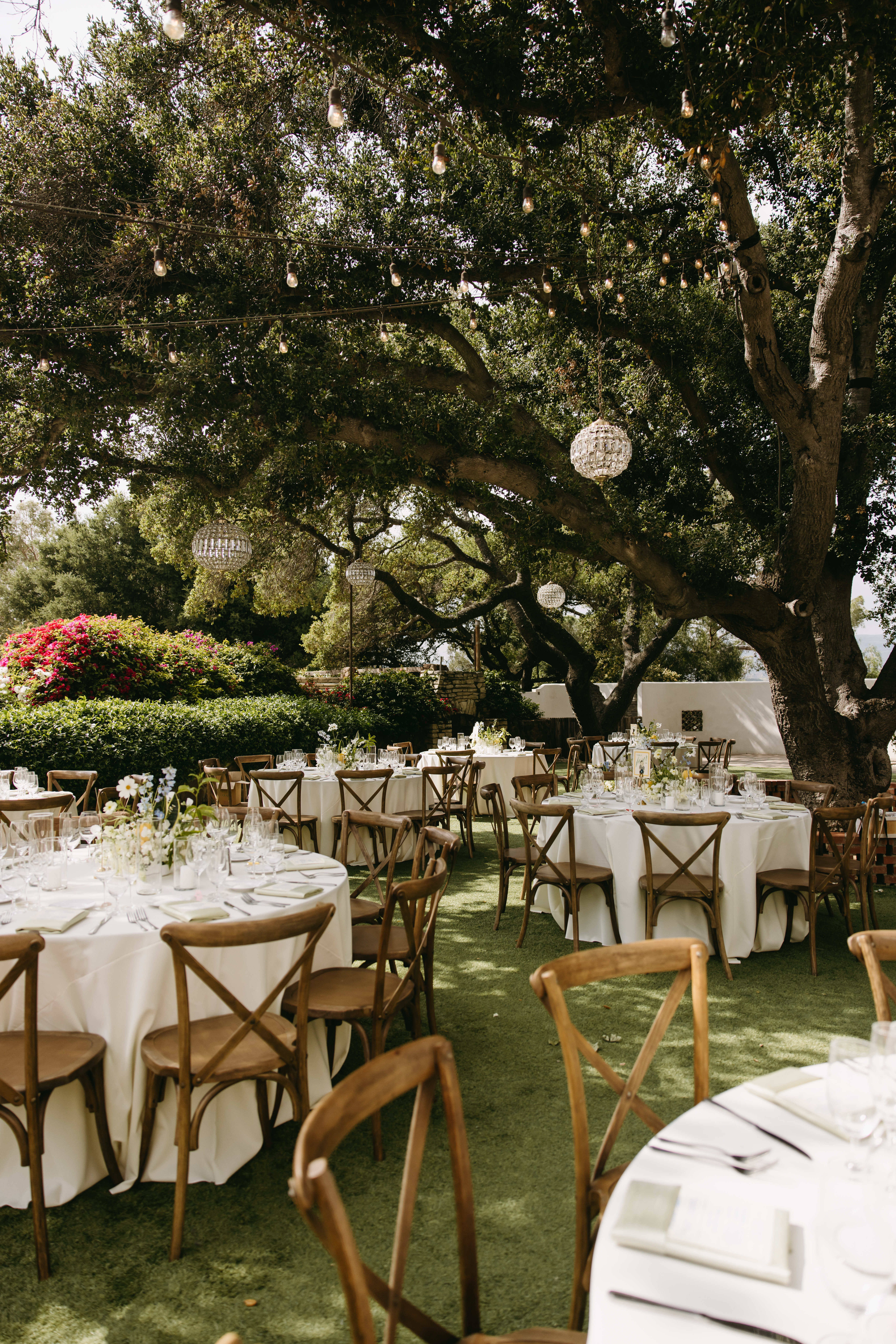 Outdoor dining area with round tables covered in white tablecloths. Wooden chairs surround the tables. Overhead, chandeliers hang from tree branches. Lush greenery is visible in the background at quail ranch