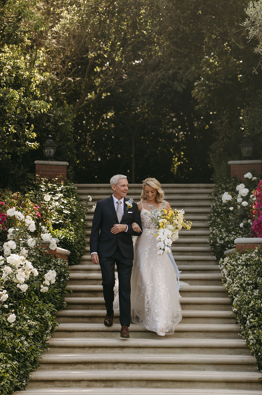 A bride in a white gown walks arm-in-arm with a man in a suit down a garden path, holding a bouquet of yellow and white flowers for a wedding at quail ranch in simi valley