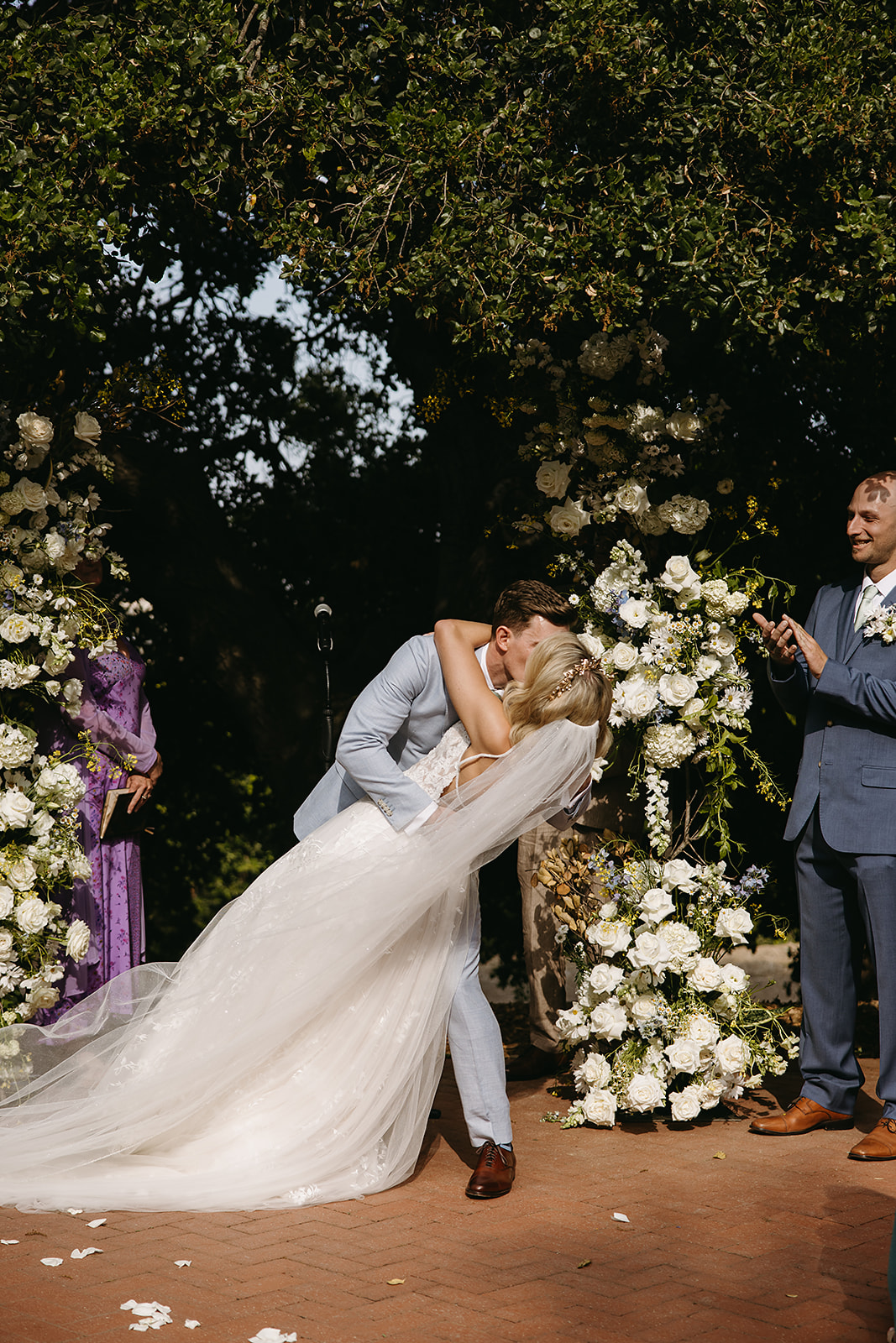 Bride and groom holding hands and smiling, walking down an outdoor aisle surrounded by flowers and guests.