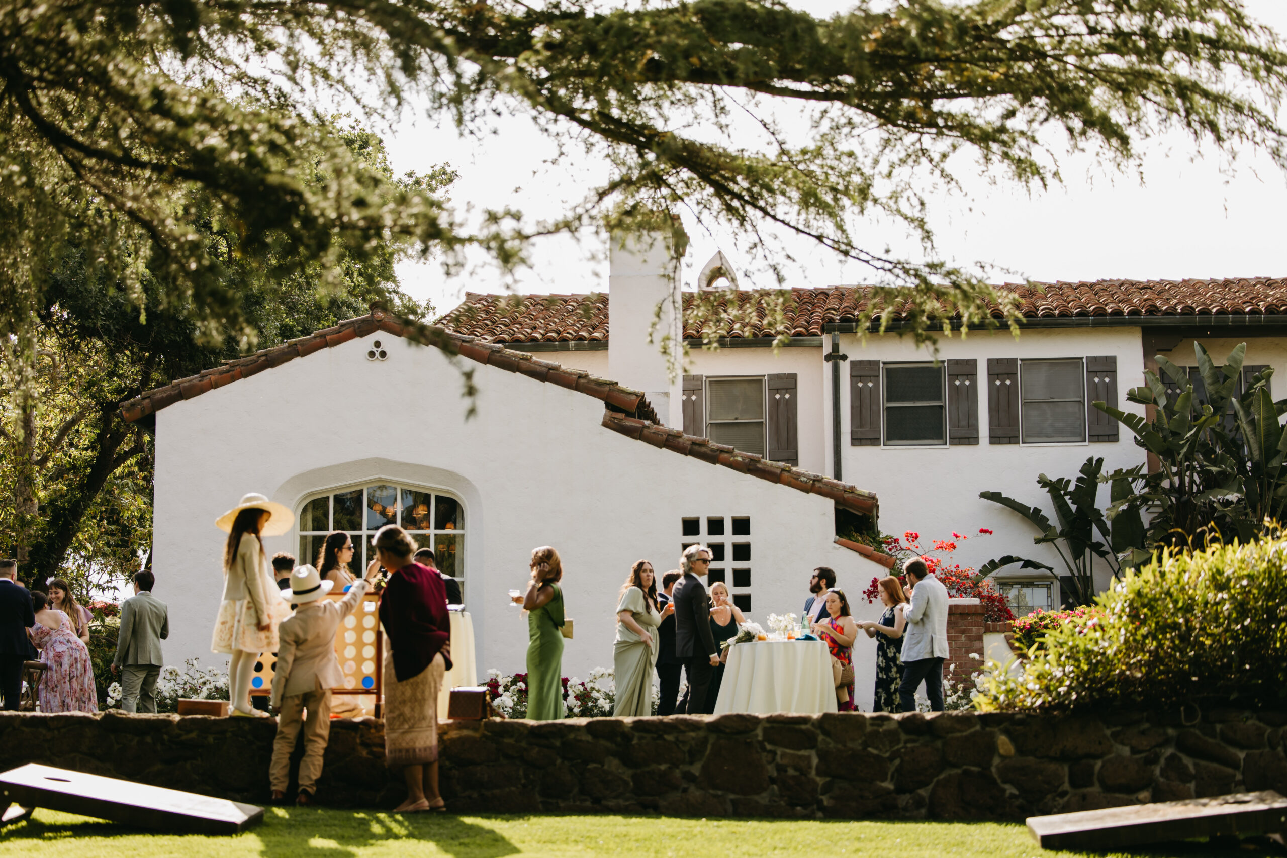 People gather outside a white, two-story house with a red-tiled roof, surrounded by trees. A lawn is in the foreground at quail ranch