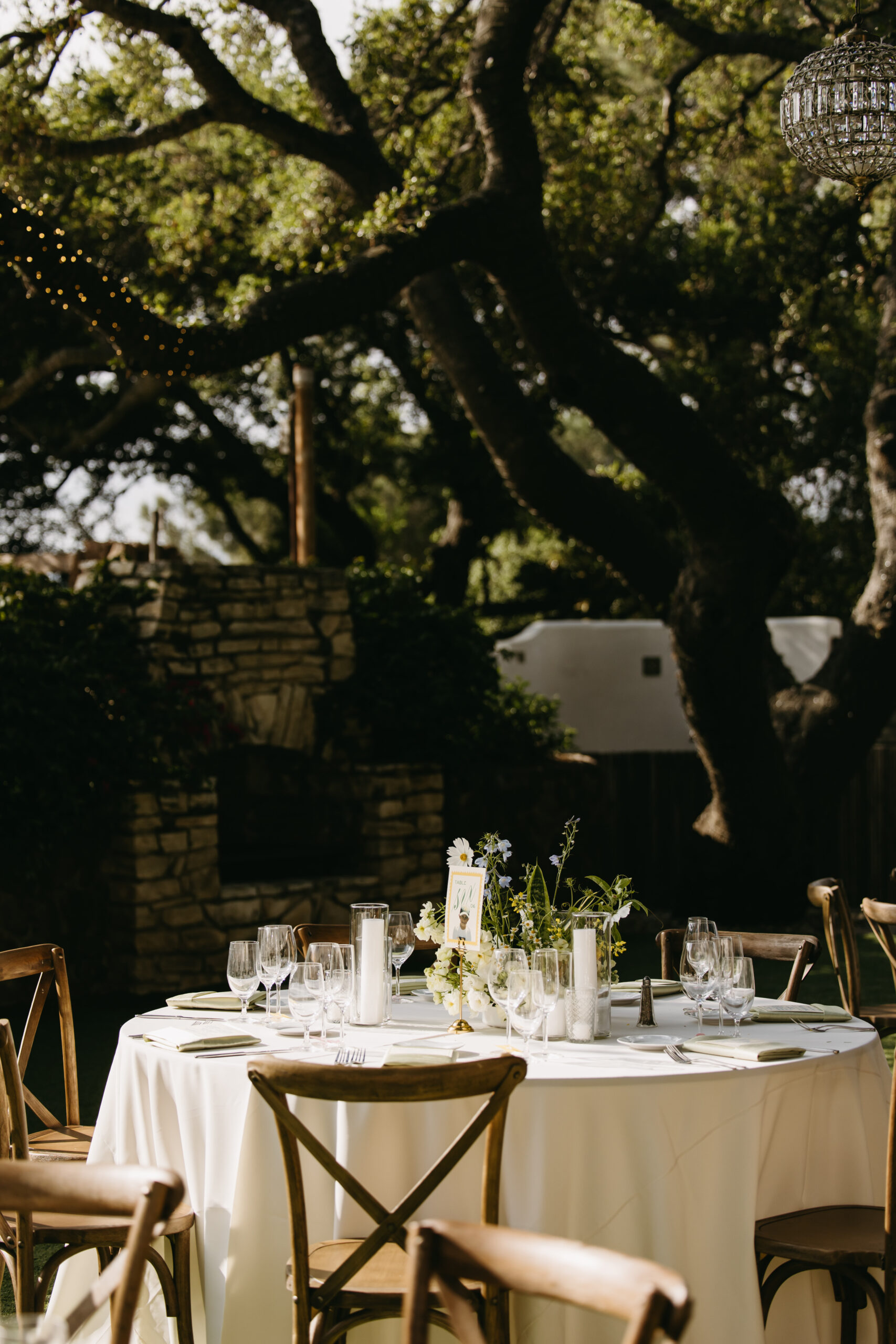 Outdoor dining area with round tables covered in white tablecloths. Wooden chairs surround the tables. Overhead, chandeliers hang from tree branches. Lush greenery is visible in the background at quail ranch