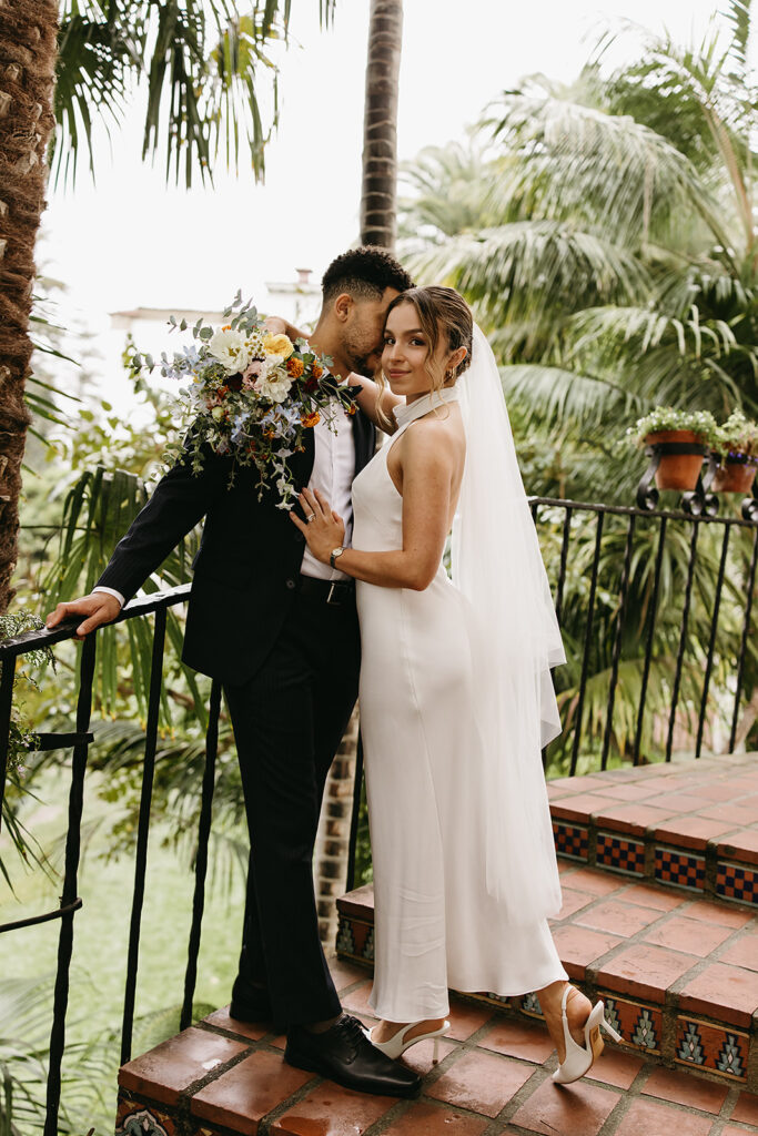 A bride and groom stand on a tiled staircase surrounded by greenery. The bride holds a bouquet and wears a white dress with a veil; the groom wears a black suit for their santa barbara elopement