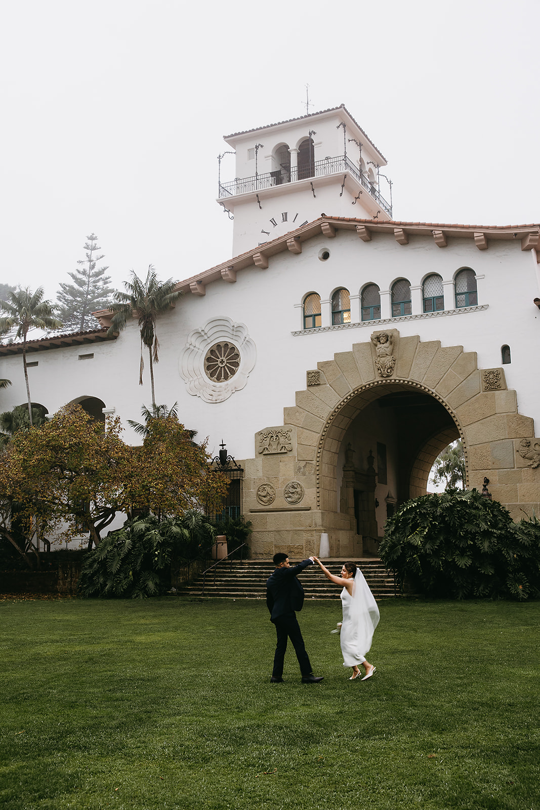 A couple dances on a grassy lawn in front of a large, historic white building with arched entryways and a clock tower for a santa barbara elopement 