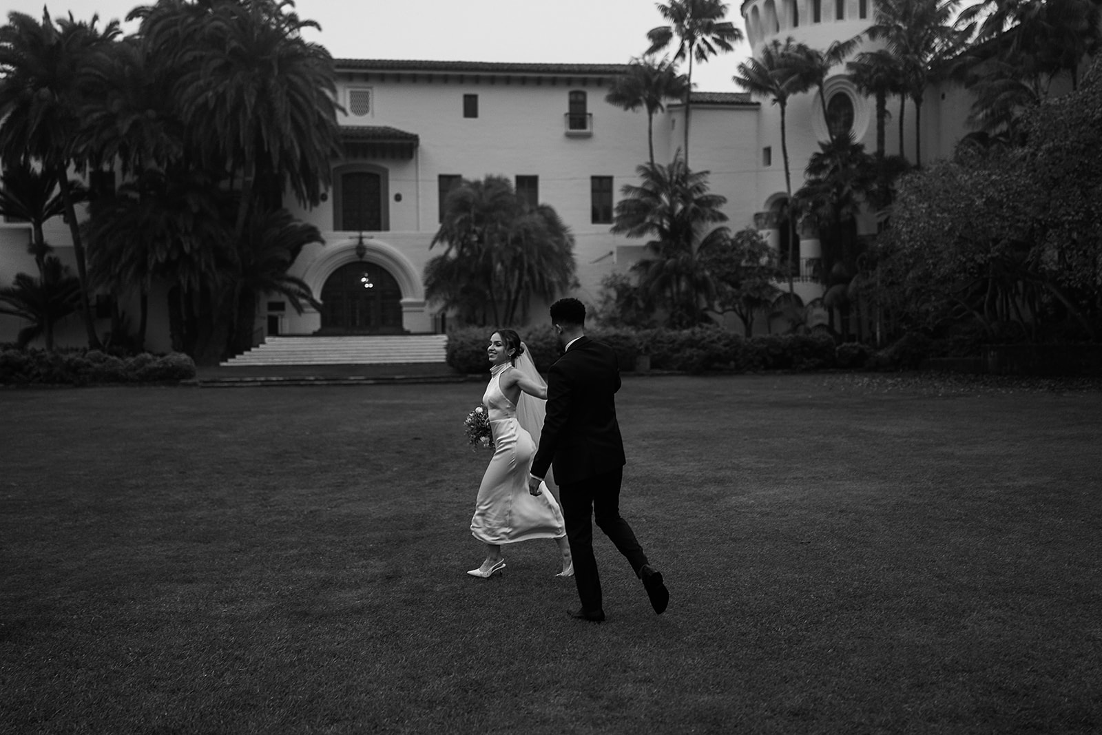 A couple dances on a grassy lawn in front of a large, historic white building with arched entryways and a clock tower for a santa barbara elopement