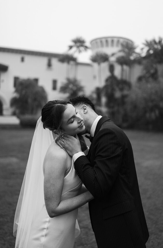 A couple in formal attire embraces outdoors on a grassy area, with the man kissing the woman's cheek. A large building and trees are in the background 