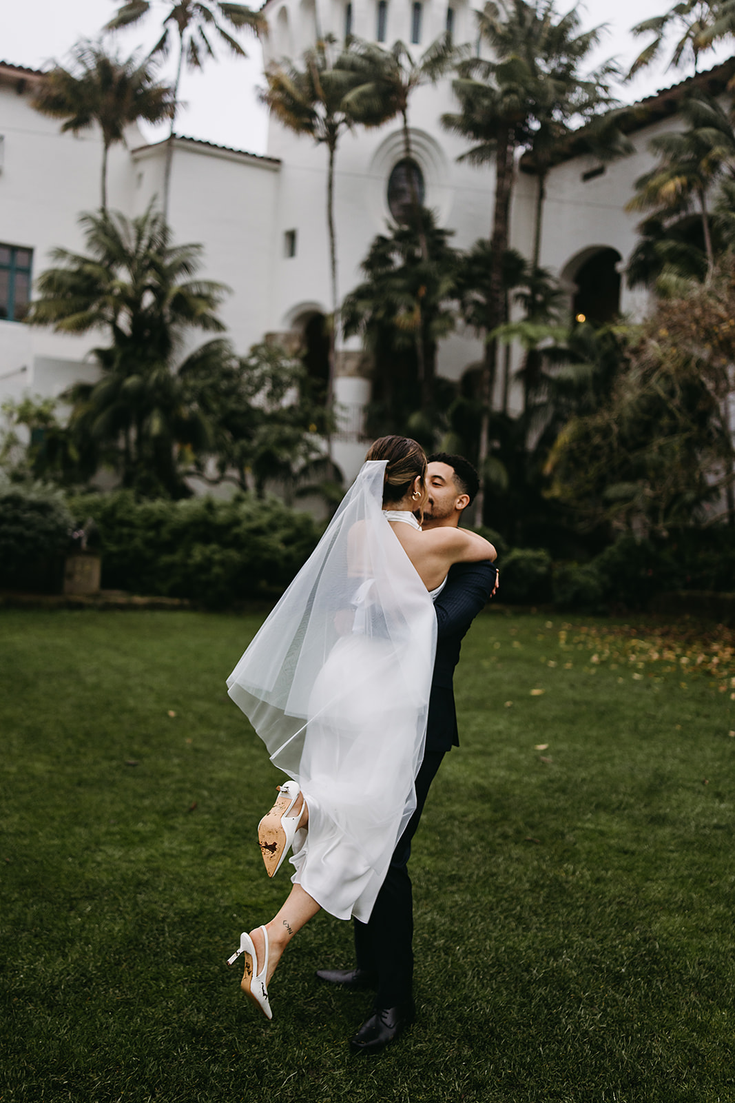 A couple dances on a grassy lawn in front of a large, historic white building with arched entryways and a clock tower for a santa barbara elopement