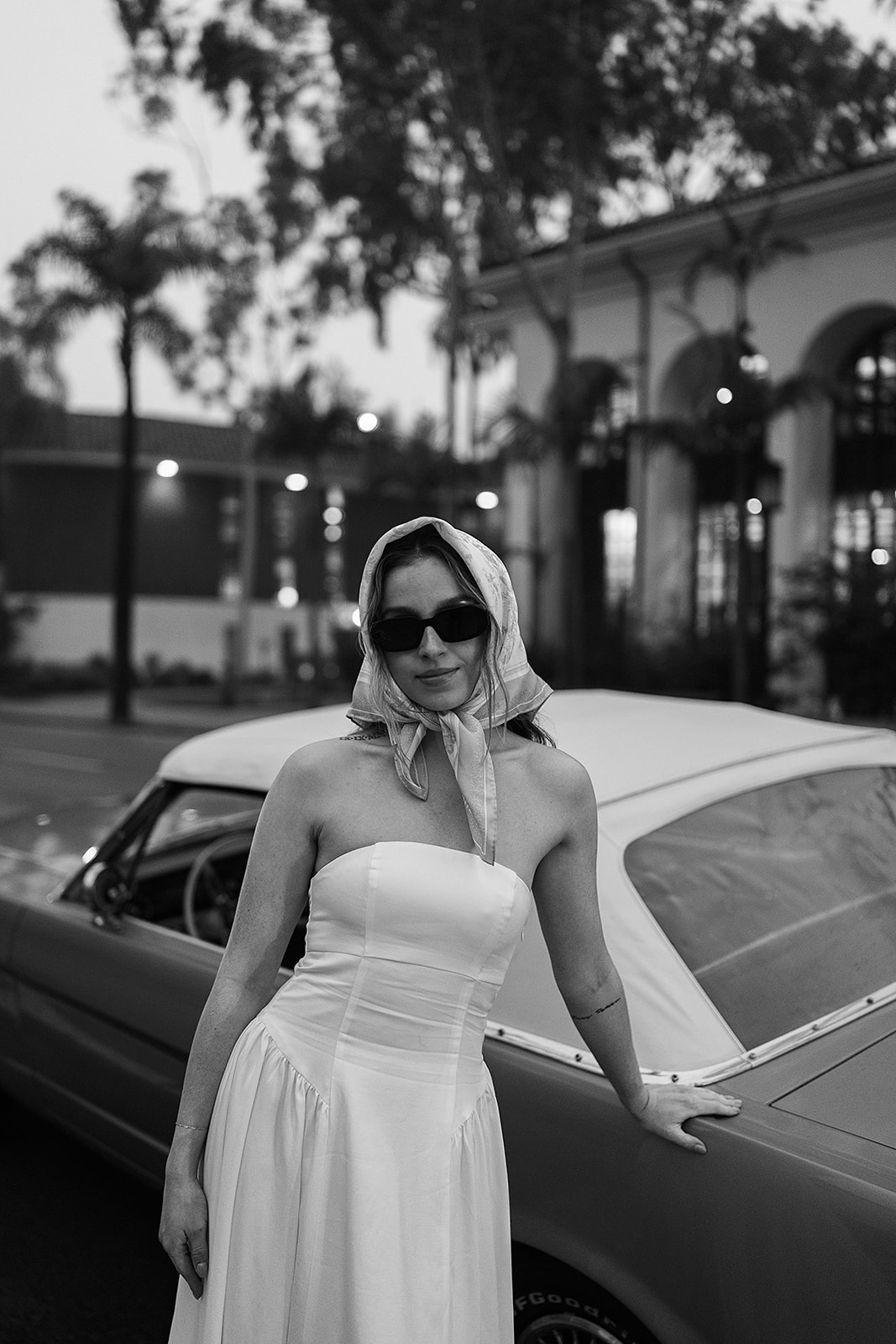 A woman in a strapless dress and headscarf stands beside a vintage car, wearing sunglasses. The scene is in black and white.