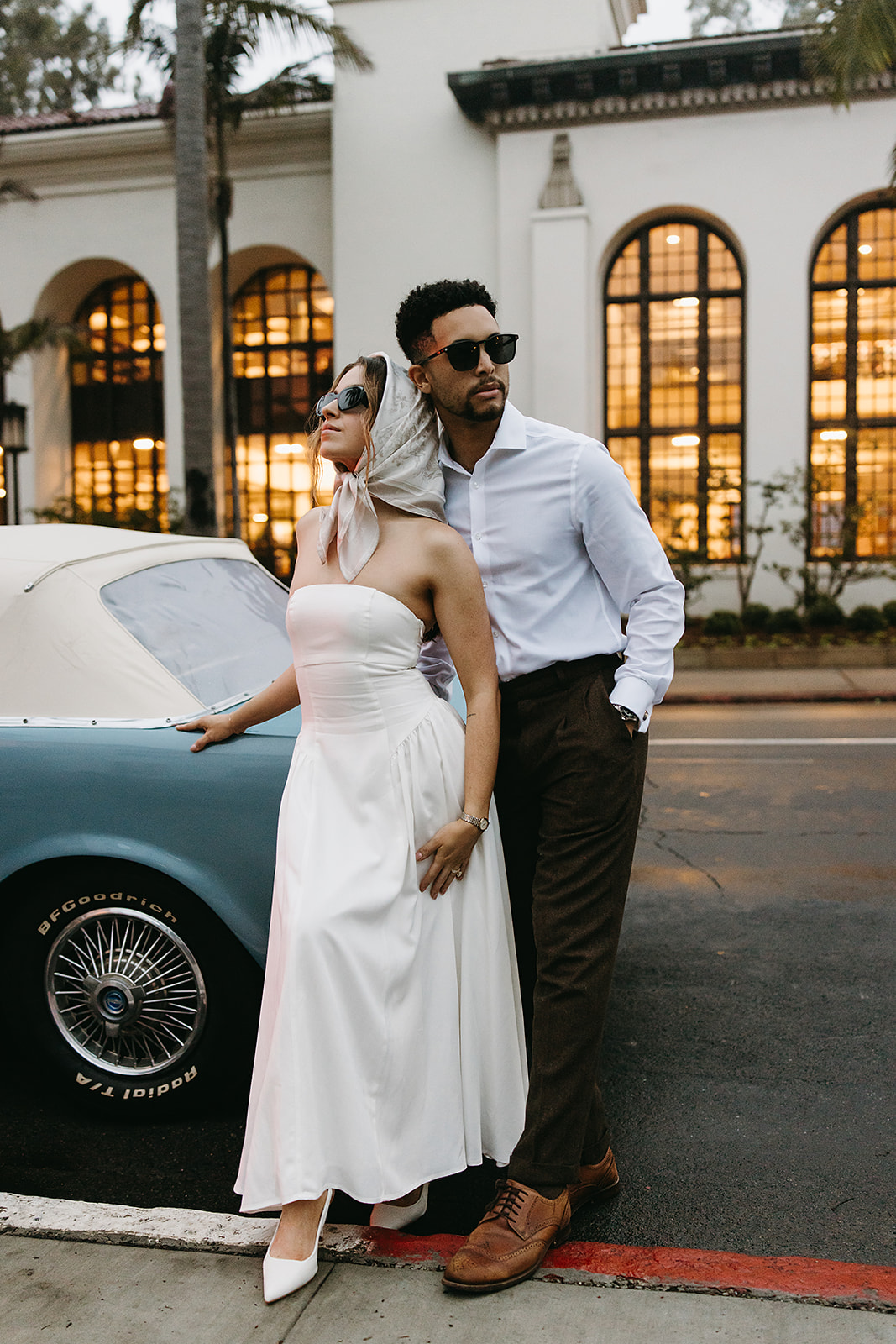 A couple in vintage attire stands by a classic car. The woman wears a white dress and sunglasses, and the man wears a white shirt and dark pants. They pose in front of a building with arched windows.