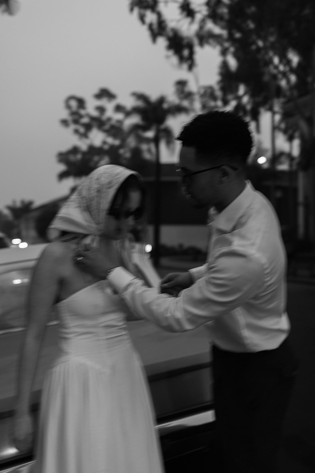 A man helps a woman adjust her scarf near a car in a blurry, black-and-white setting. Trees are visible in the background.