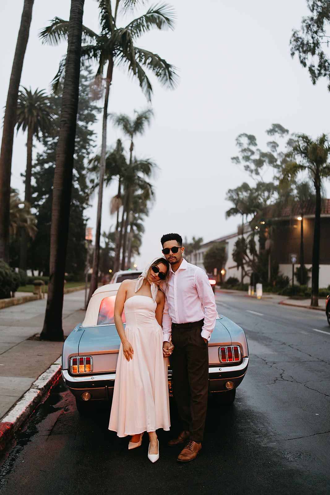 A couple in vintage attire stands by a classic car. The woman wears a white dress and sunglasses, and the man wears a white shirt and dark pants. They pose in front of a building with arched windows for their santa barbara elopement