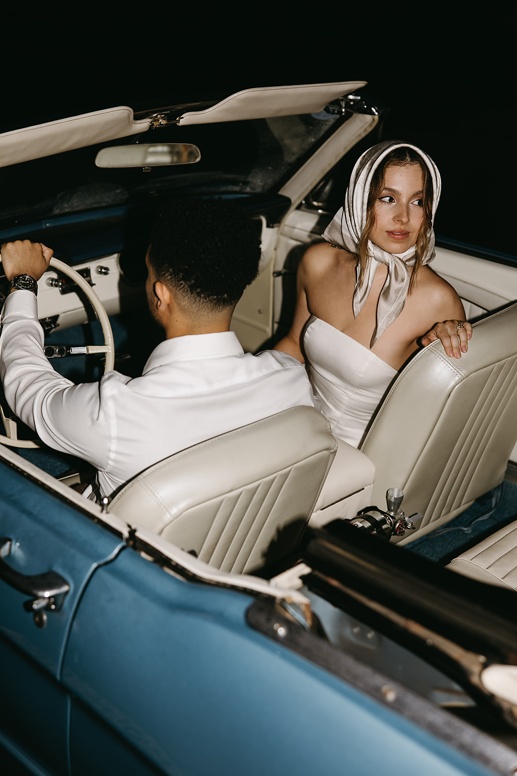 Bride and groom sit in a vintage convertible, the bride holding a bouquet. The lighting is dim, casting a warm glow inside the car for their santa barbara elopment