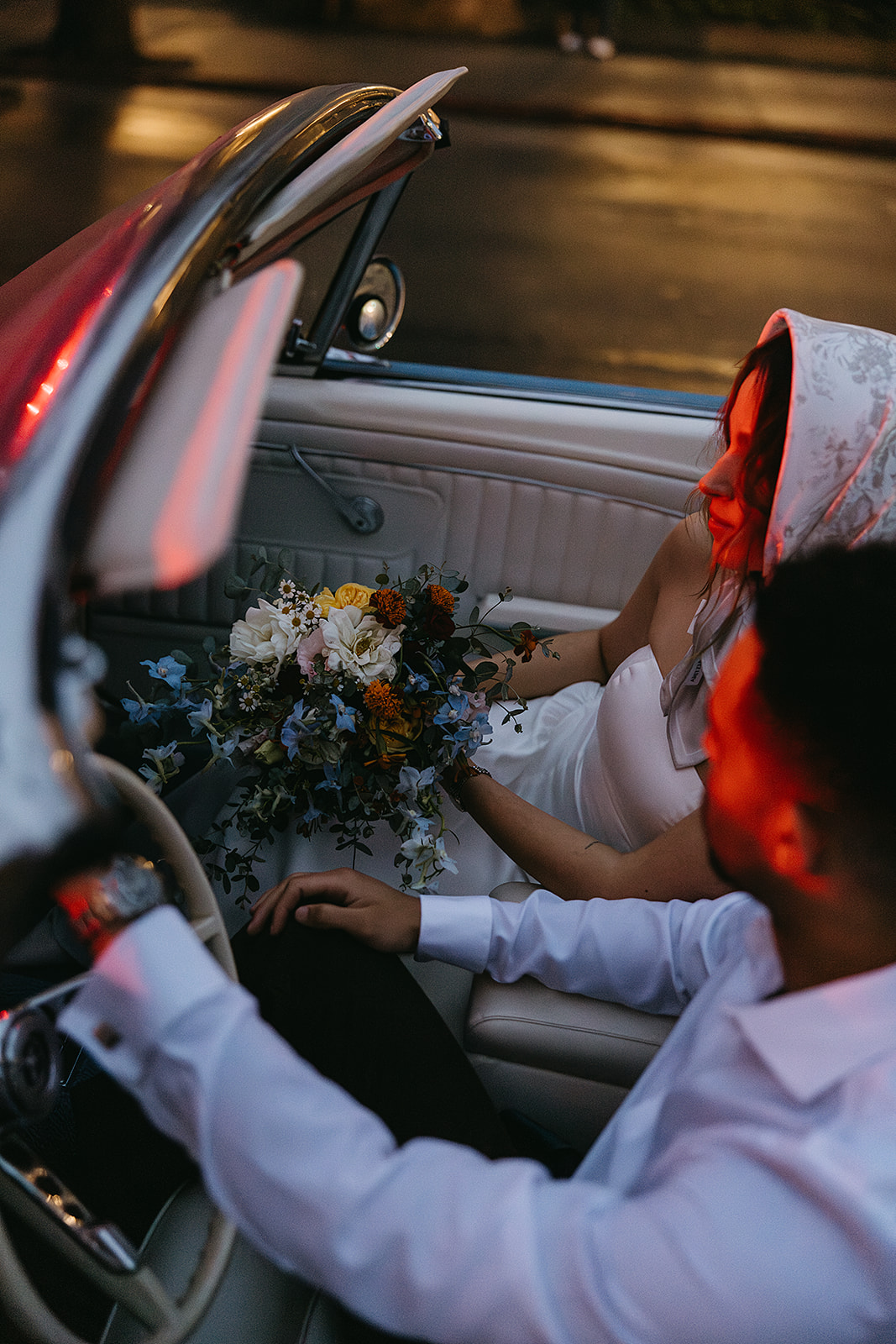 Bride and groom sit in a vintage convertible, the bride holding a bouquet. The lighting is dim, casting a warm glow inside the car for their santa barbara elopment