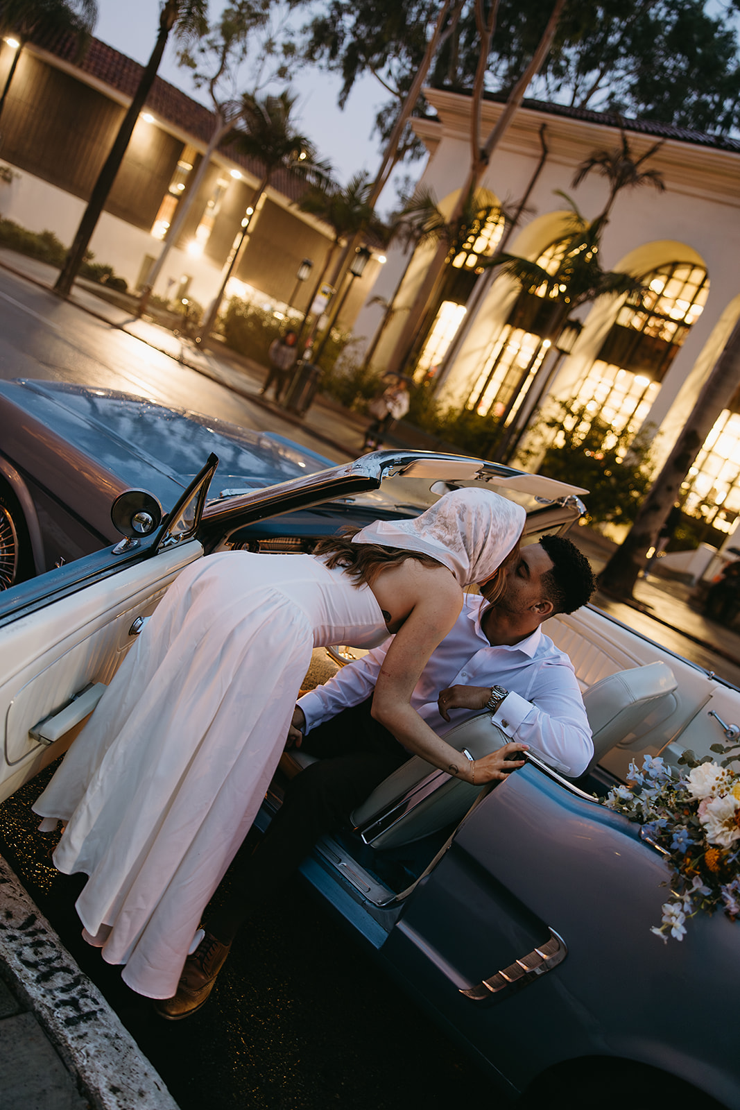 A couple shares a kiss beside a convertible car, with a bouquet on the back seat. They're dressed in wedding attire, and the scene is set against a backdrop of buildings and trees for their santa barbara elopement