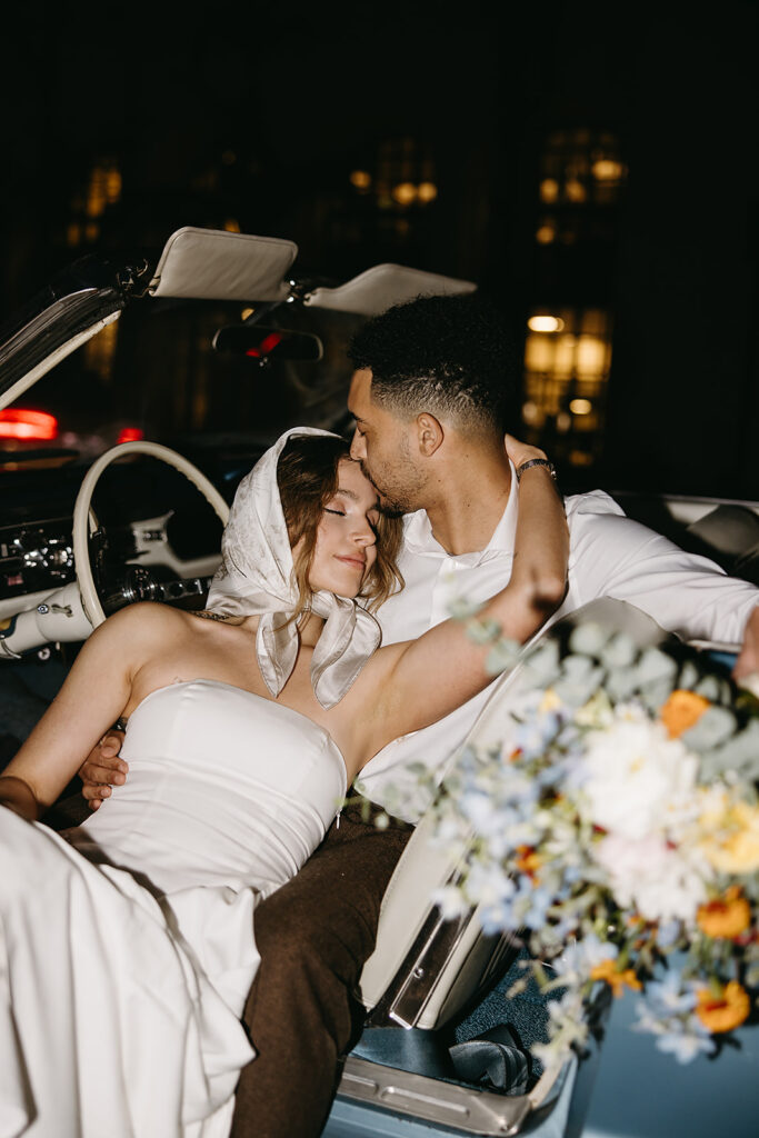 A couple in formal attire leans against a vintage blue convertible at night. at their santa barbara elopement