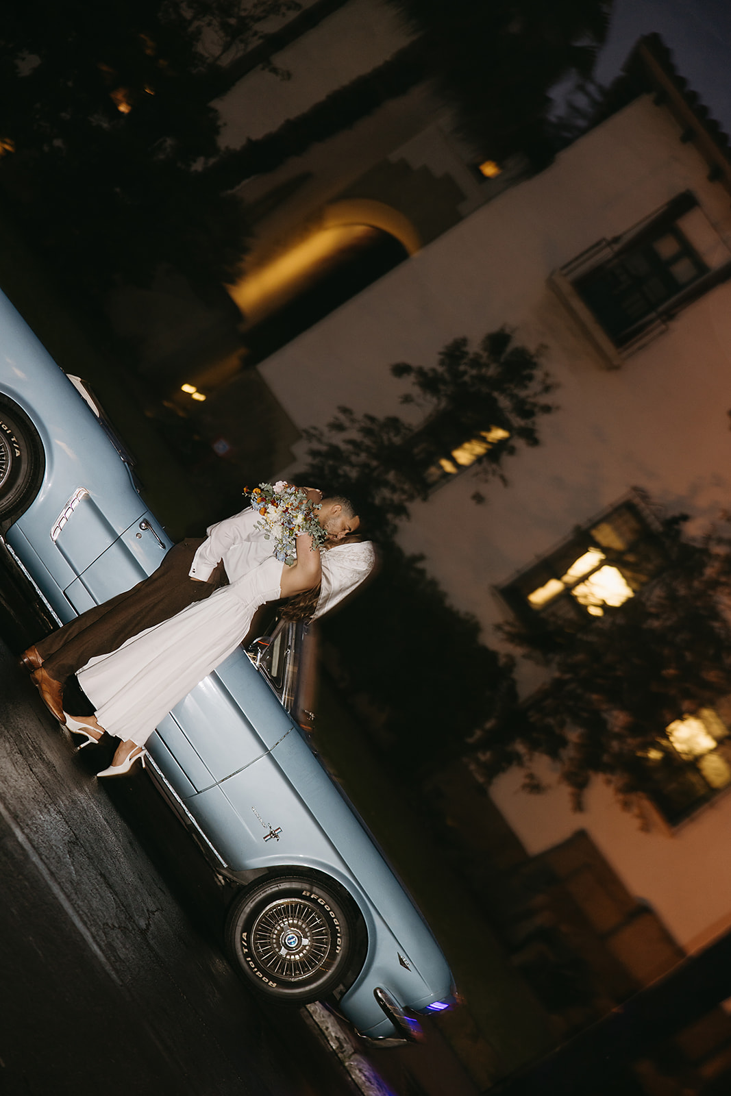 A couple in formal attire leans against a vintage blue convertible at night. at their santa barbara elopement