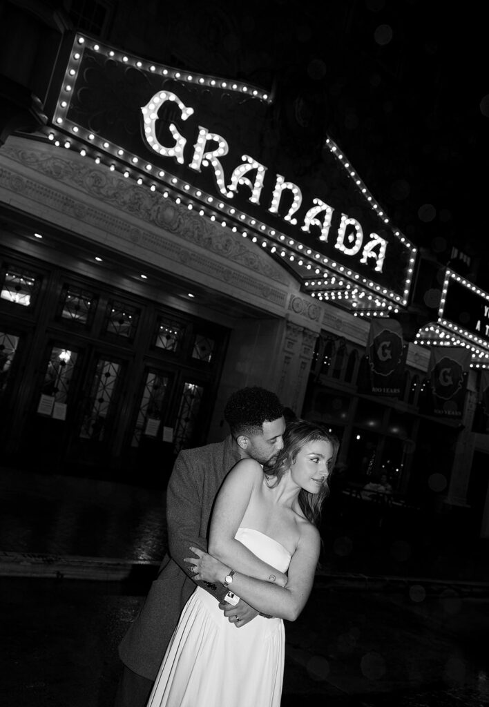 A couple embraces in front of the Granada theater at night, with bright marquee lights above them.