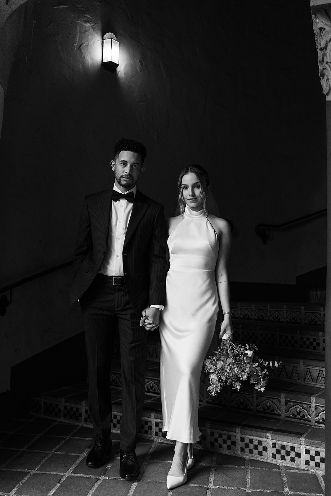 A couple stands on a tiled staircase. The man wears a suit, and the woman is in a white dress holding a bouquet. The scene is lit by a wall sconce for their santa barbara elopement 