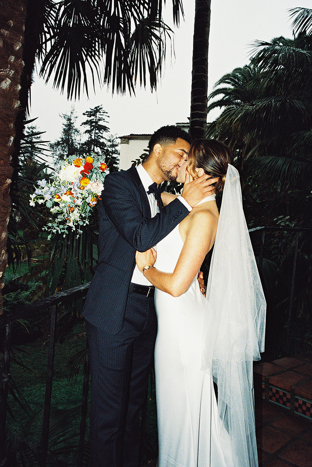 A bride and groom stand on a tiled staircase surrounded by greenery. The bride holds a bouquet and wears a white dress with a veil; the groom wears a black suit for their santa barbara elopement