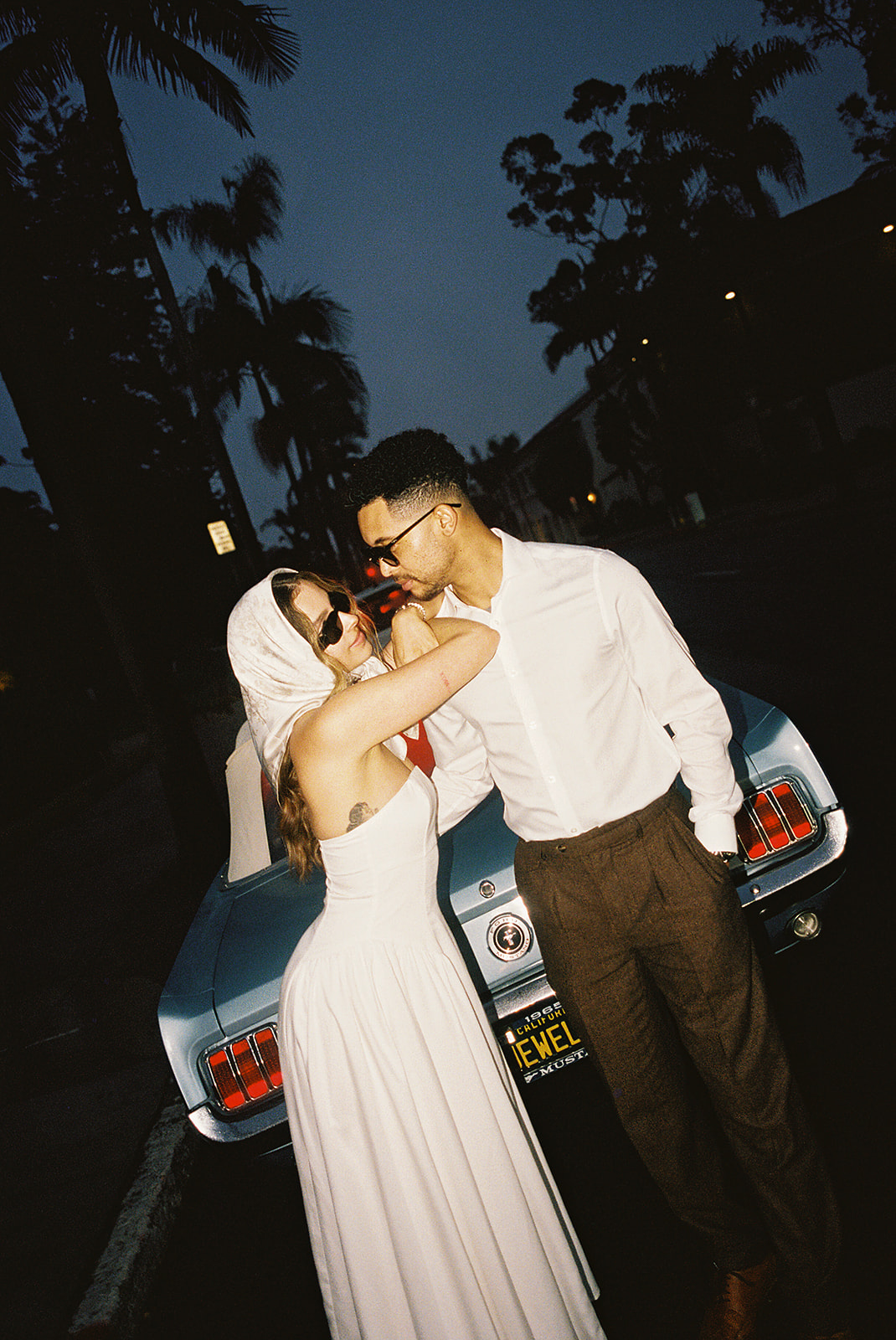 A couple in vintage attire stands by a classic car. The woman wears a white dress and sunglasses, and the man wears a white shirt and dark pants. They pose in front of a building with arched windows for their santa barbara elopement