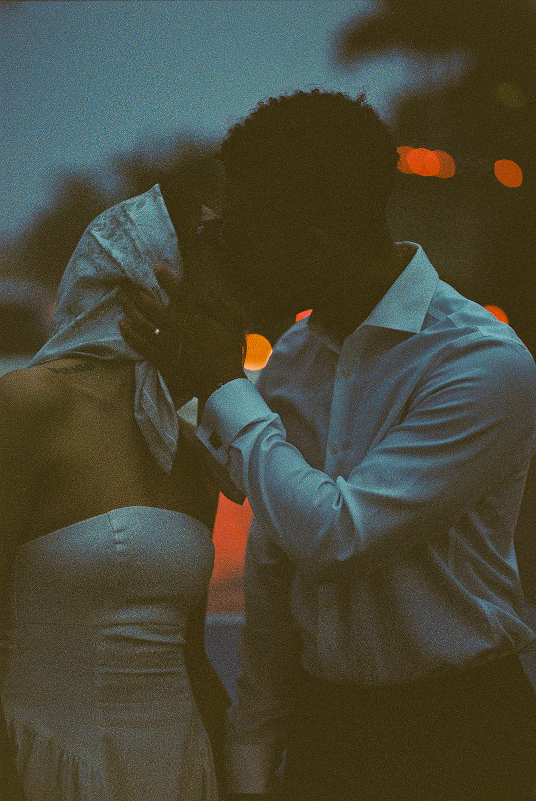 A couple kisses outdoors at dusk, illuminated by soft streetlights. The person on the left wears a strapless dress and headscarf, and the person on the right is in a button-up shirt.