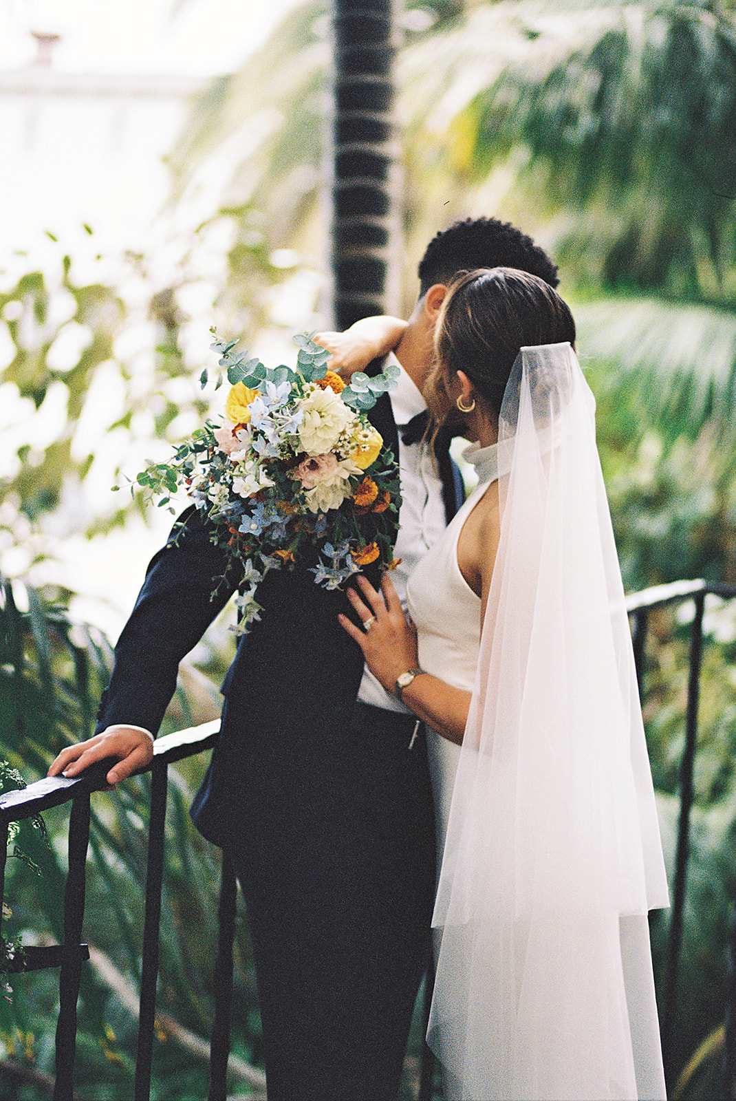 A bride and groom stand on a tiled staircase surrounded by greenery. The bride holds a bouquet and wears a white dress with a veil; the groom wears a black suit for their santa barbara elopement