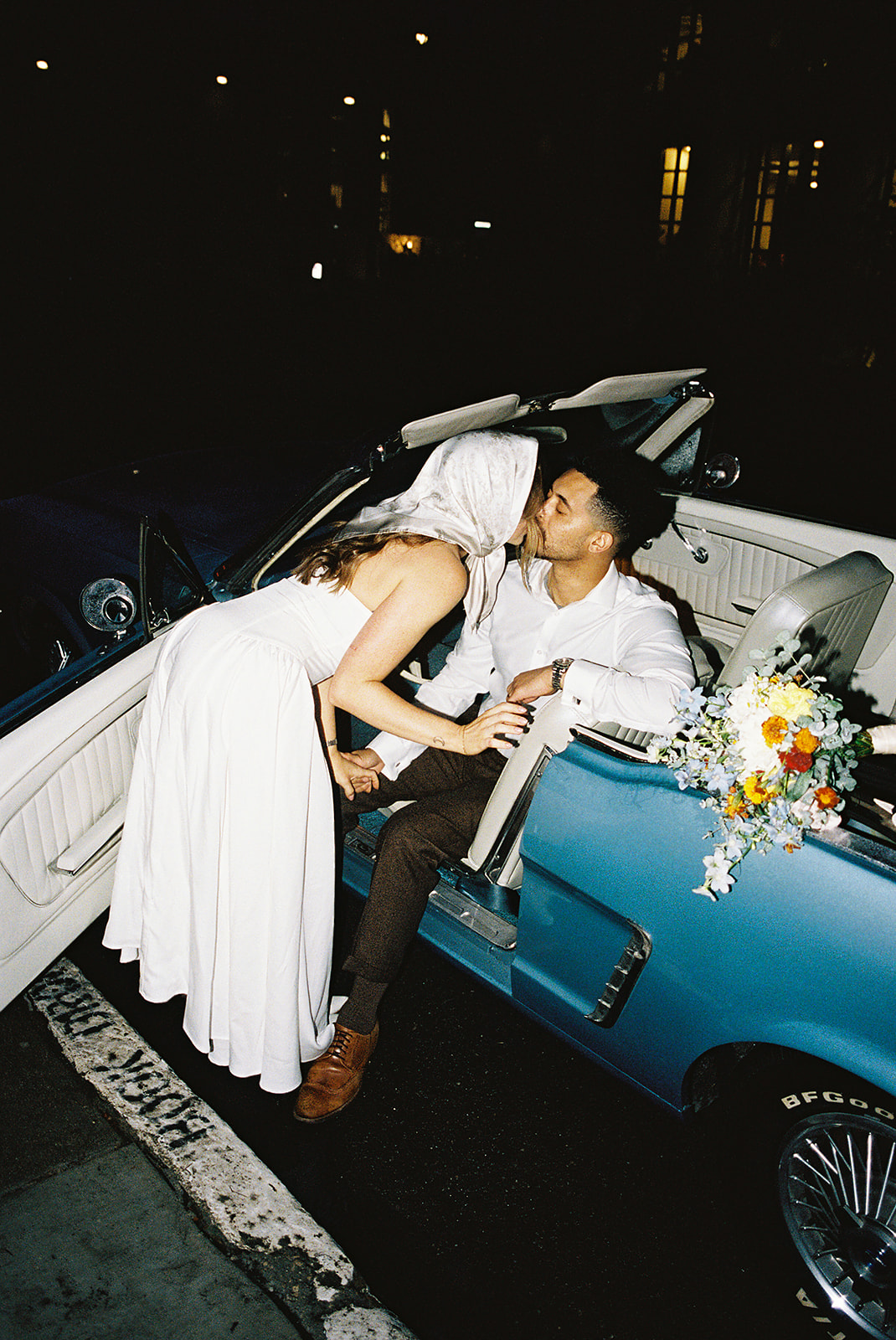 A couple kisses outdoors at dusk, illuminated by soft streetlights. The person on the left wears a strapless dress and headscarf, and the person on the right is in a button-up shirt.