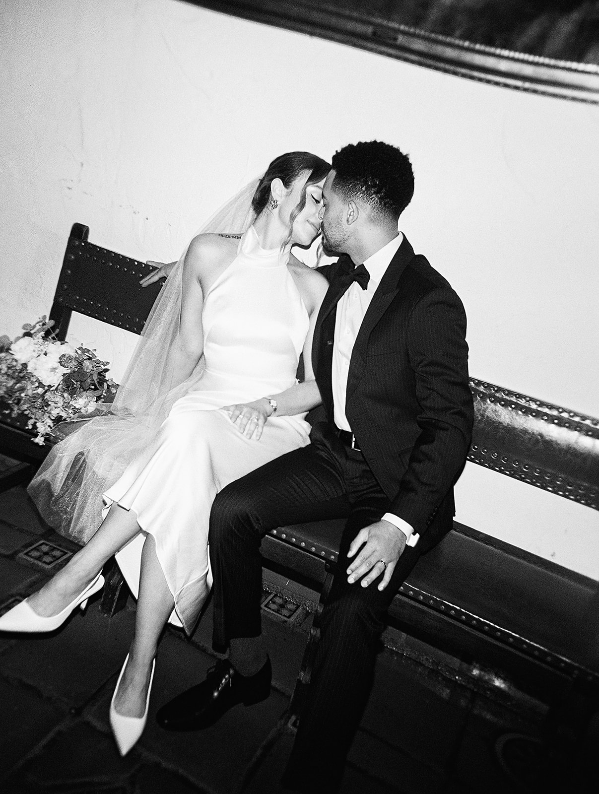 A couple in formal attire embraces outdoors on a grassy area, with the man kissing the woman's cheek. A large building and trees are in the background