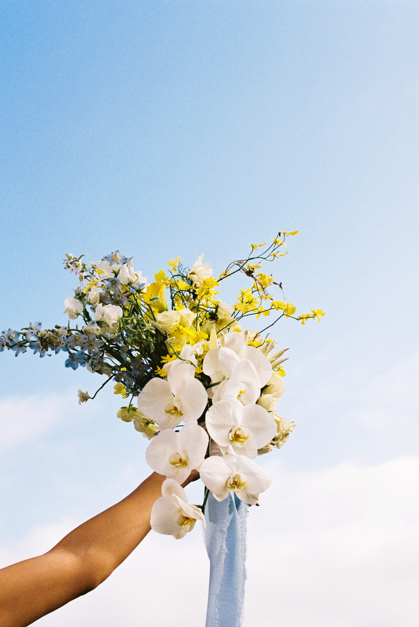 A hand holds a bouquet of white orchids and yellow flowers against a clear blue sky.