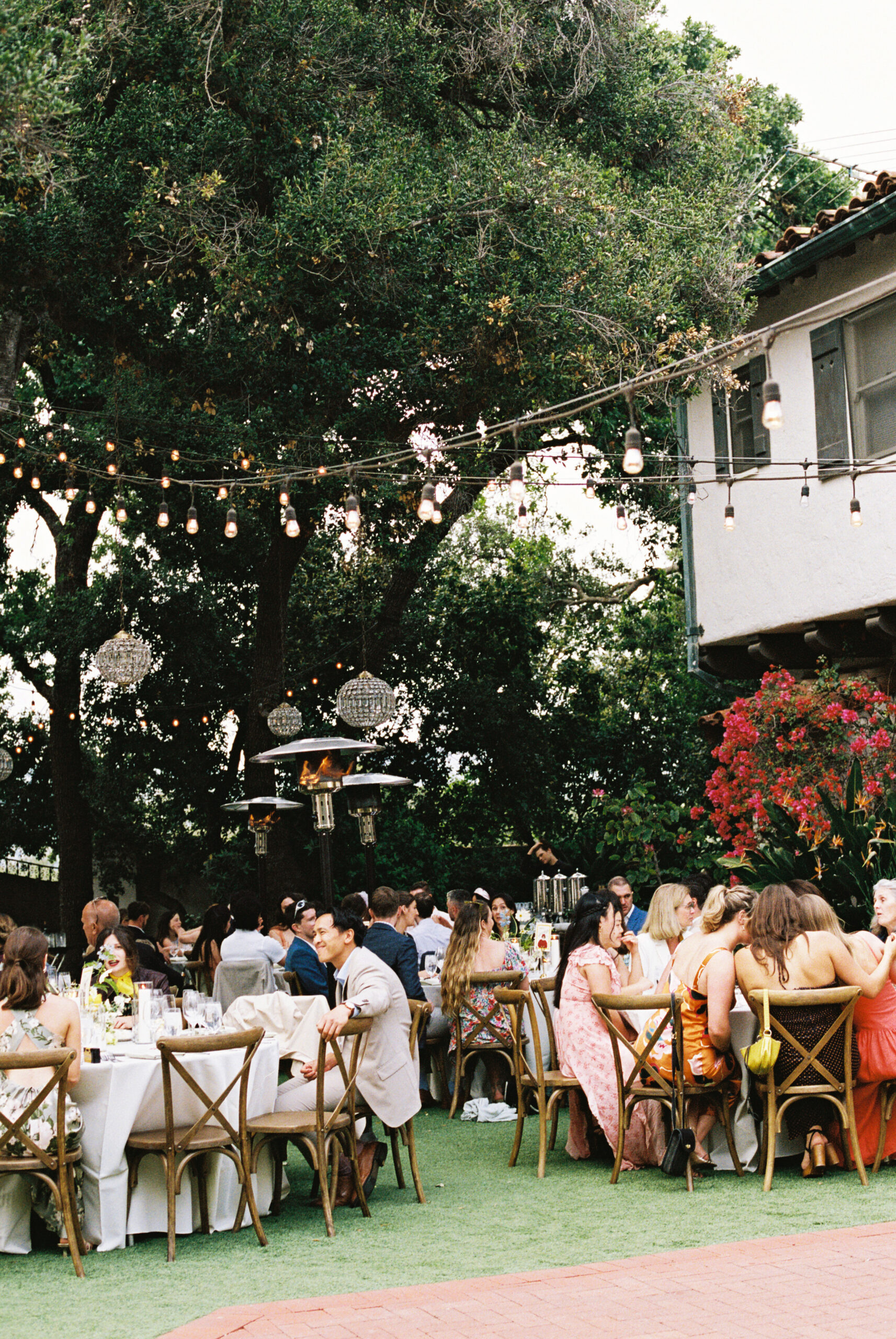 Outdoor dining area with round tables covered in white tablecloths. Wooden chairs surround the tables. Overhead, chandeliers hang from tree branches. Lush greenery is visible in the background.