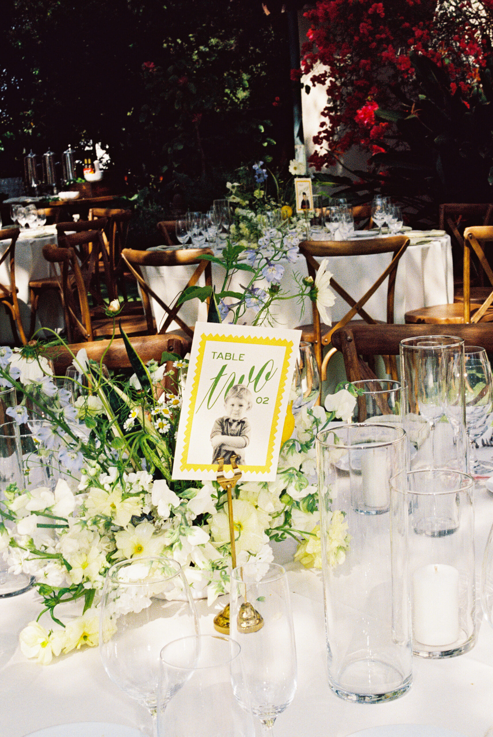 Outdoor dining area with round tables covered in white tablecloths. Wooden chairs surround the tables. Overhead, chandeliers hang from tree branches. Lush greenery is visible in the background.