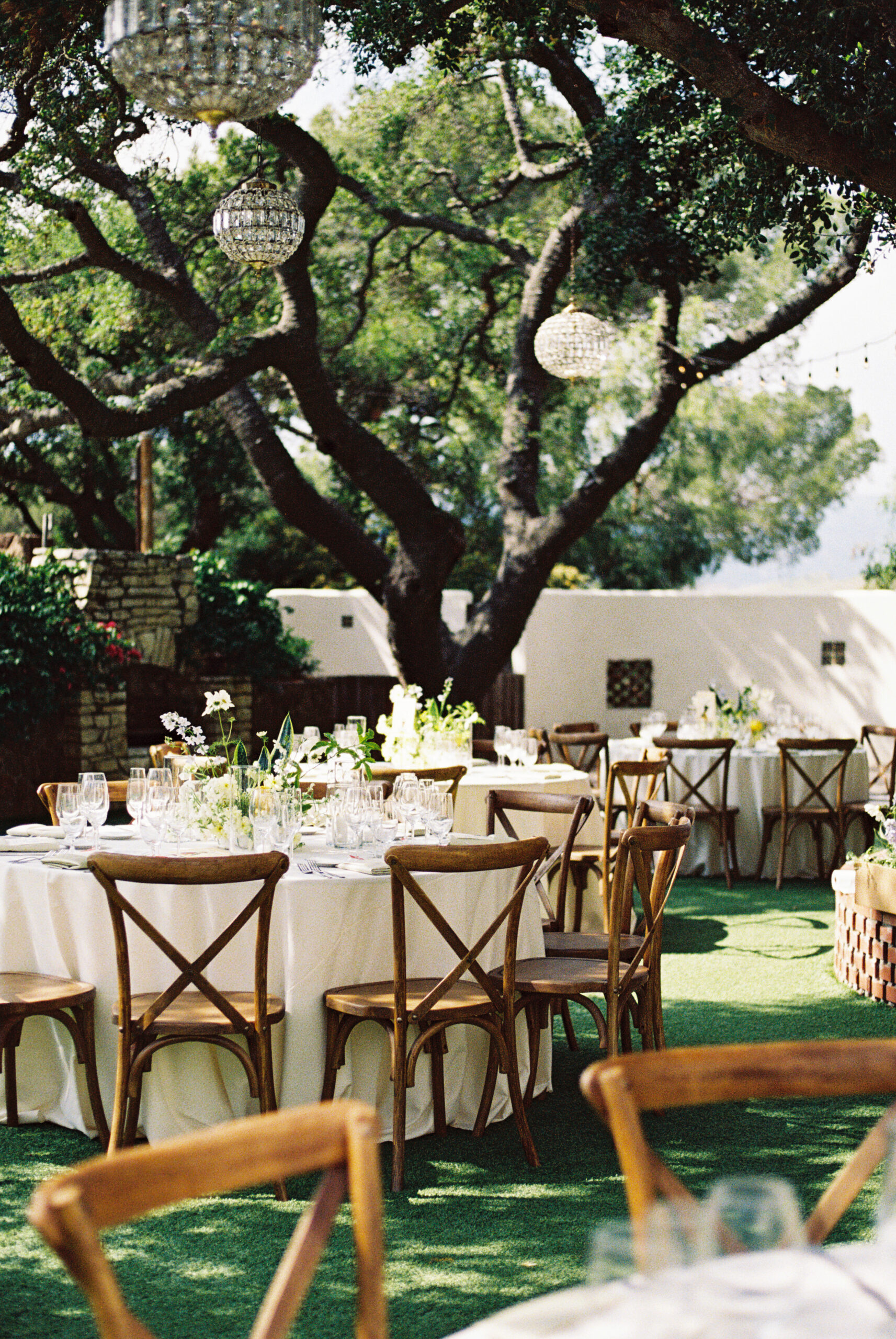 Outdoor dining area with round tables covered in white tablecloths. Wooden chairs surround the tables. Overhead, chandeliers hang from tree branches. Lush greenery is visible in the background.