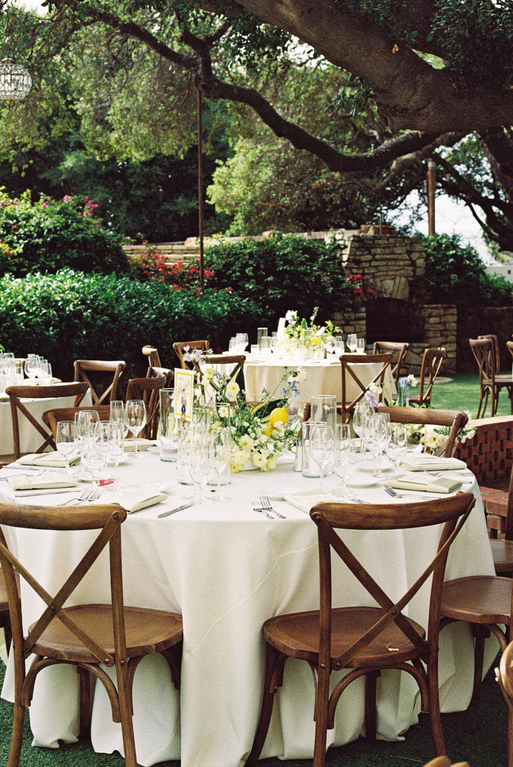 Outdoor dining area with round tables covered in white tablecloths. Wooden chairs surround the tables. Overhead, chandeliers hang from tree branches. Lush greenery is visible in the background.