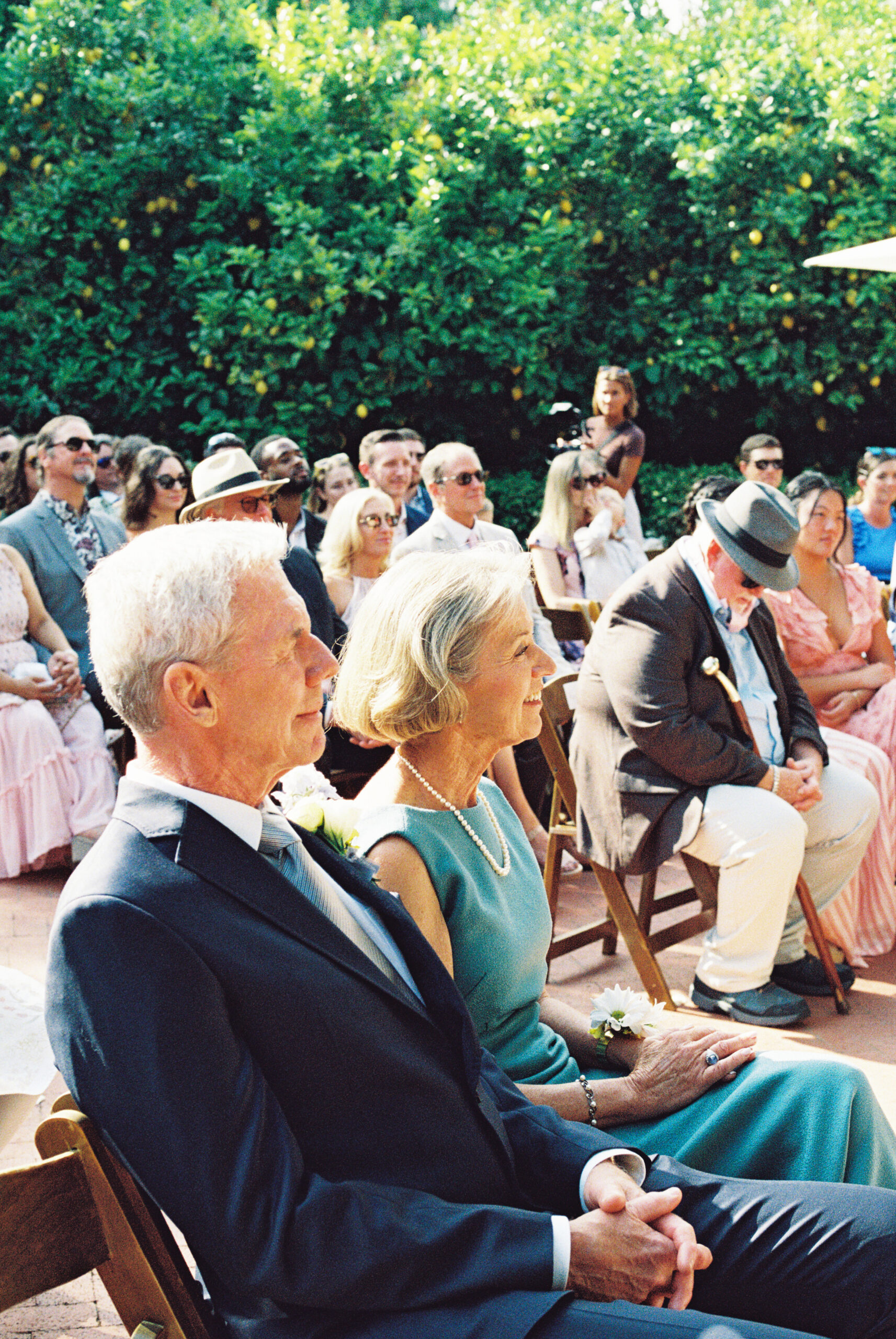 A group of people seated outdoors at a wedding ceremony, with two older individuals in the foreground wearing formal attire.