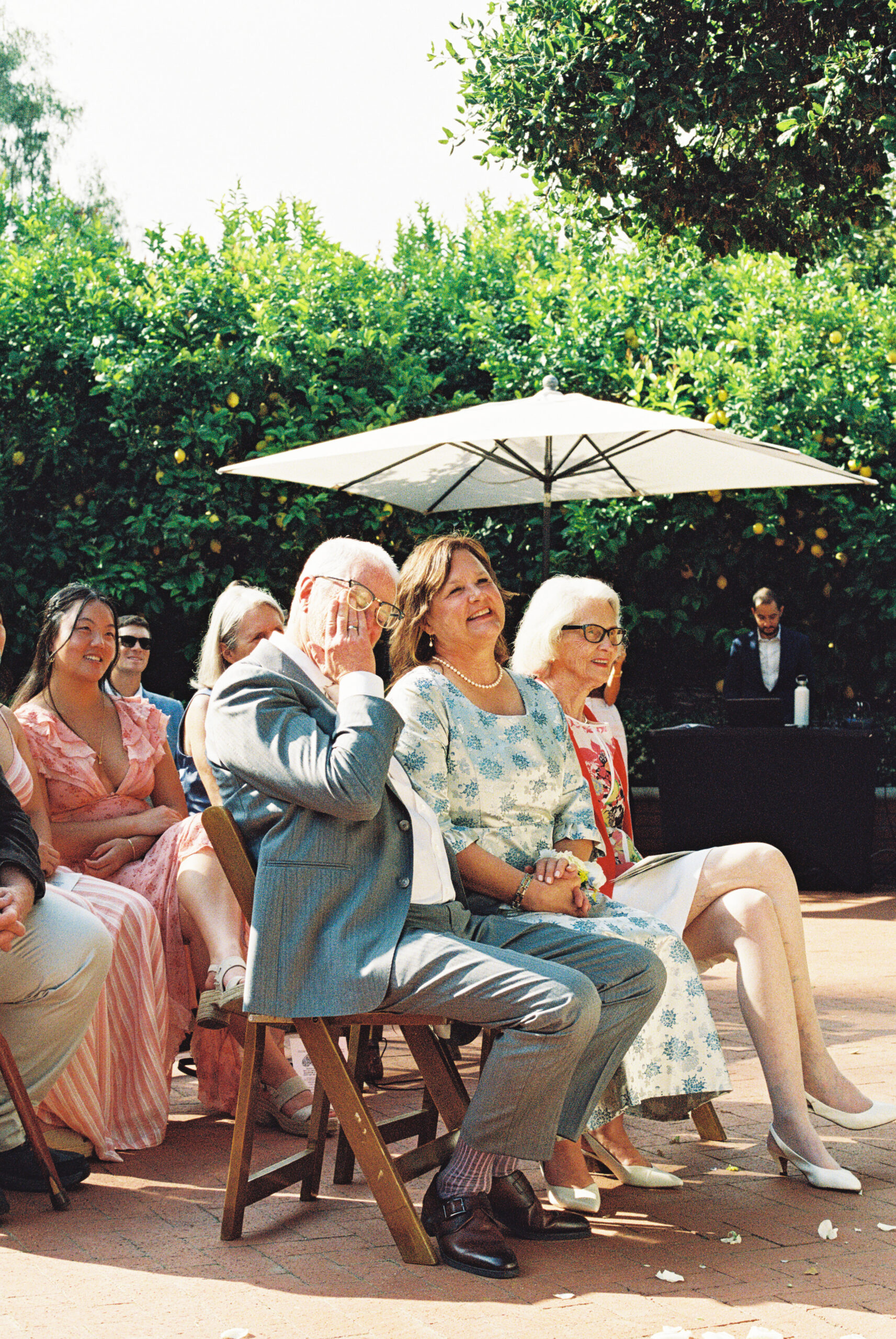 A group of people seated outdoors at a wedding ceremony, with two older individuals in the foreground wearing formal attire.