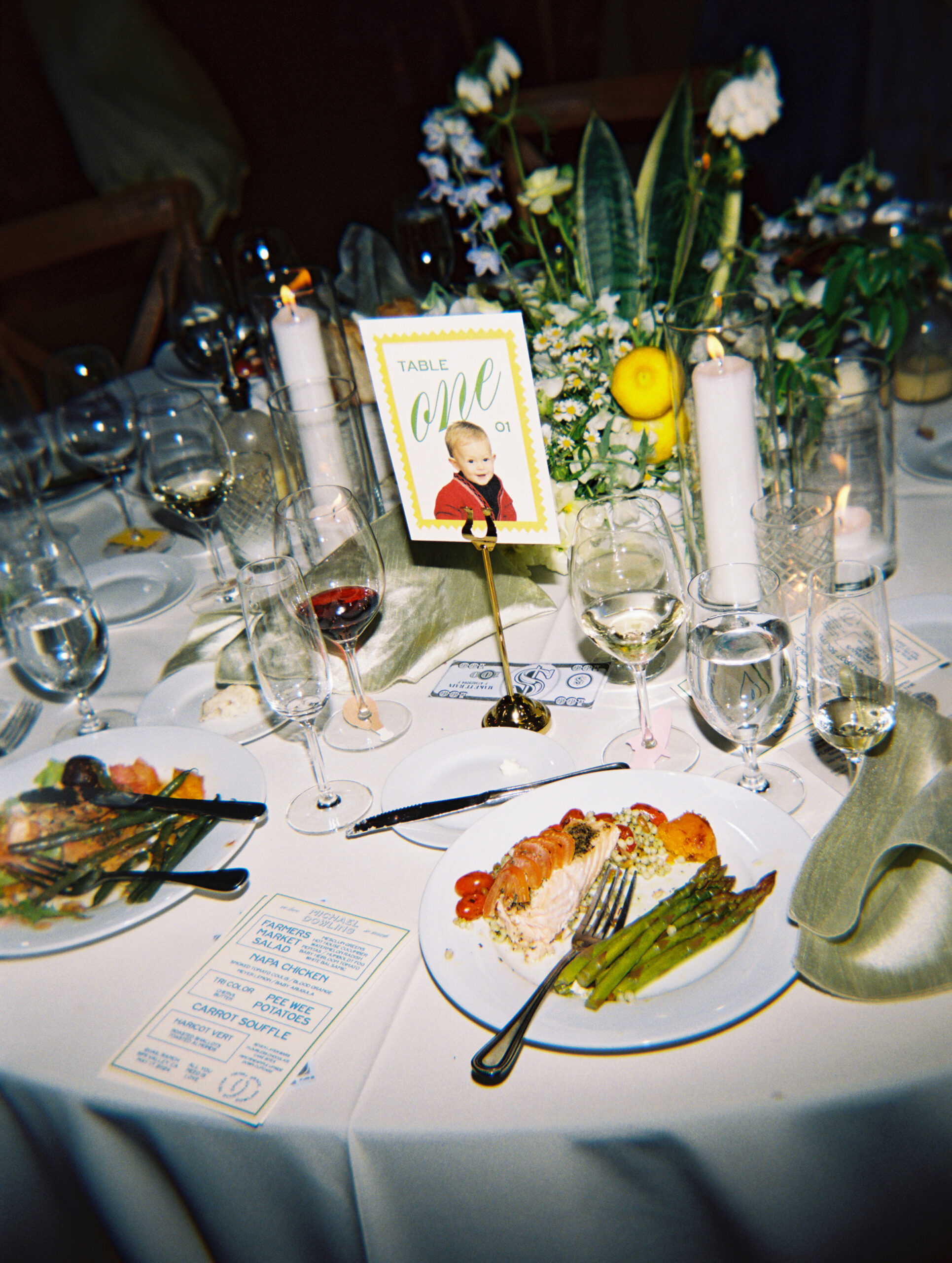 A formal table setting with plates of food, wine glasses, and a centerpiece with flowers. A menu and a photo card are placed on the table.