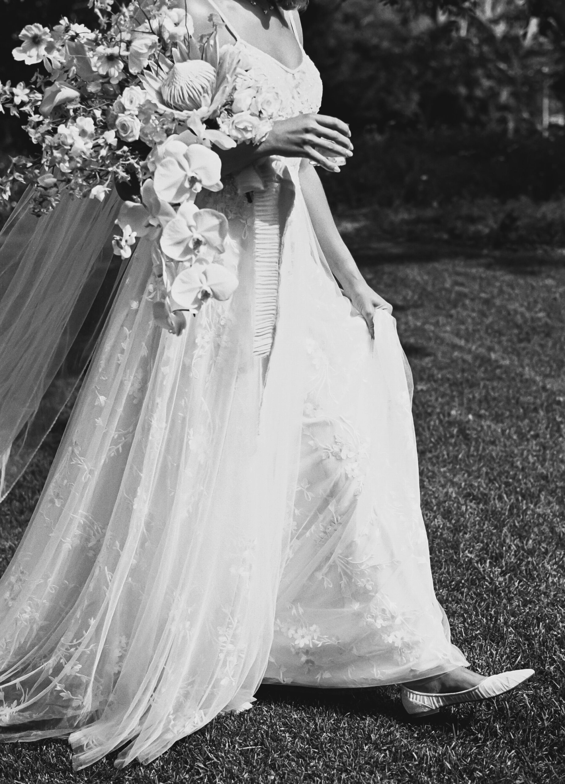 Bride walking on grass in a lace wedding dress, holding a bouquet of flowers. Black and white image.