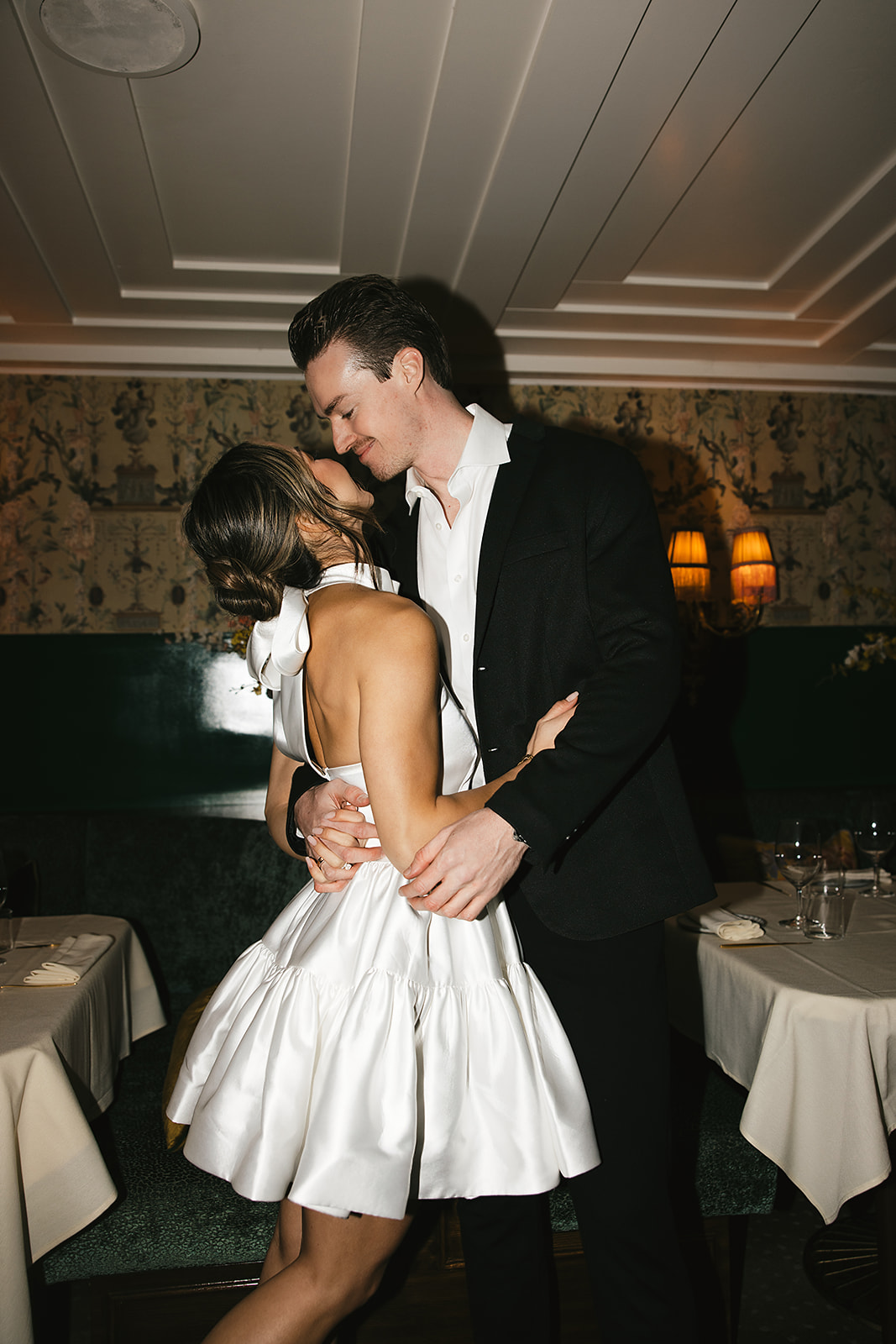 A couple in formal attire shares a dance in a restaurant with white tablecloths and dim lighting.