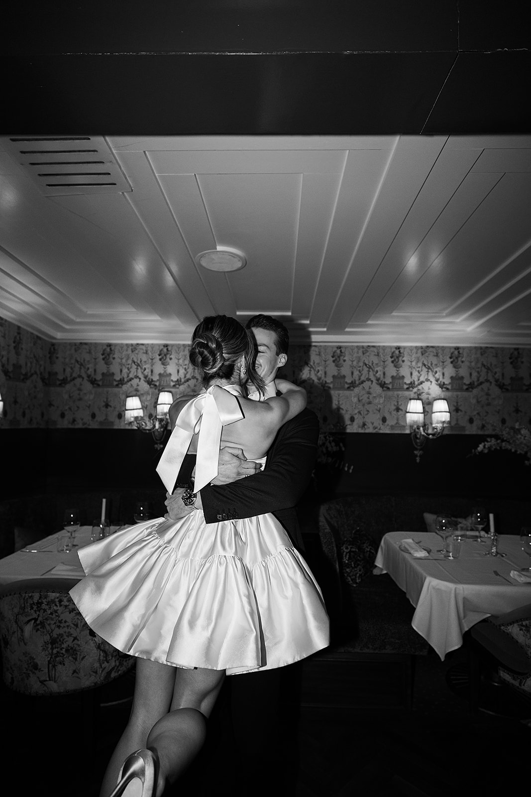 A couple in formal attire shares a dance in a restaurant with white tablecloths and dim lighting.