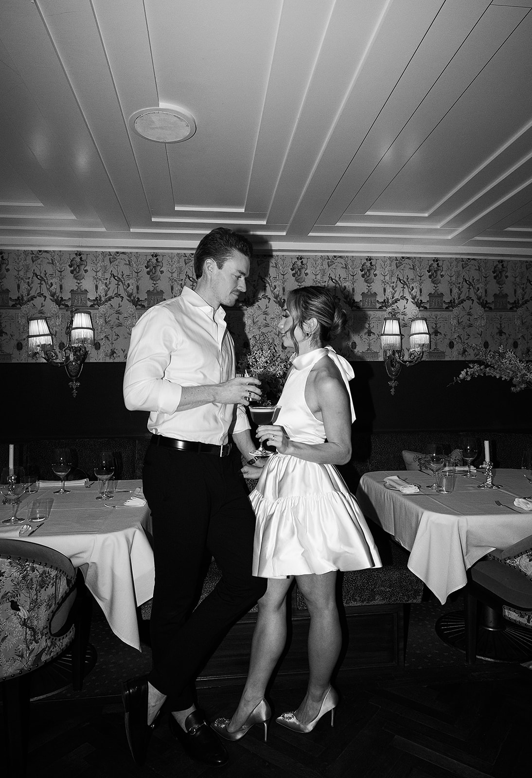 A couple in formal attire shares a dance in a restaurant with white tablecloths and dim lighting.