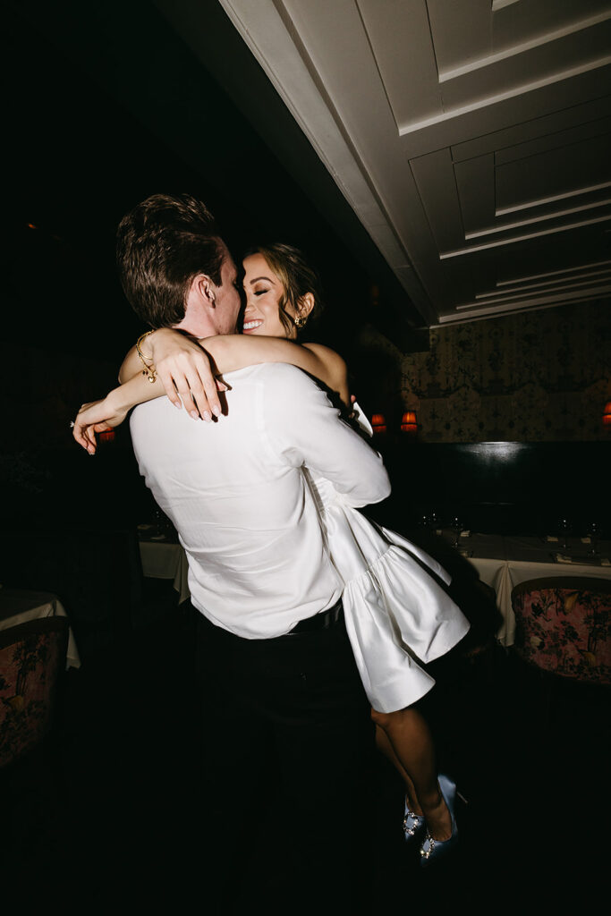 A couple in formal attire shares a dance in a restaurant with white tablecloths and dim lighting.