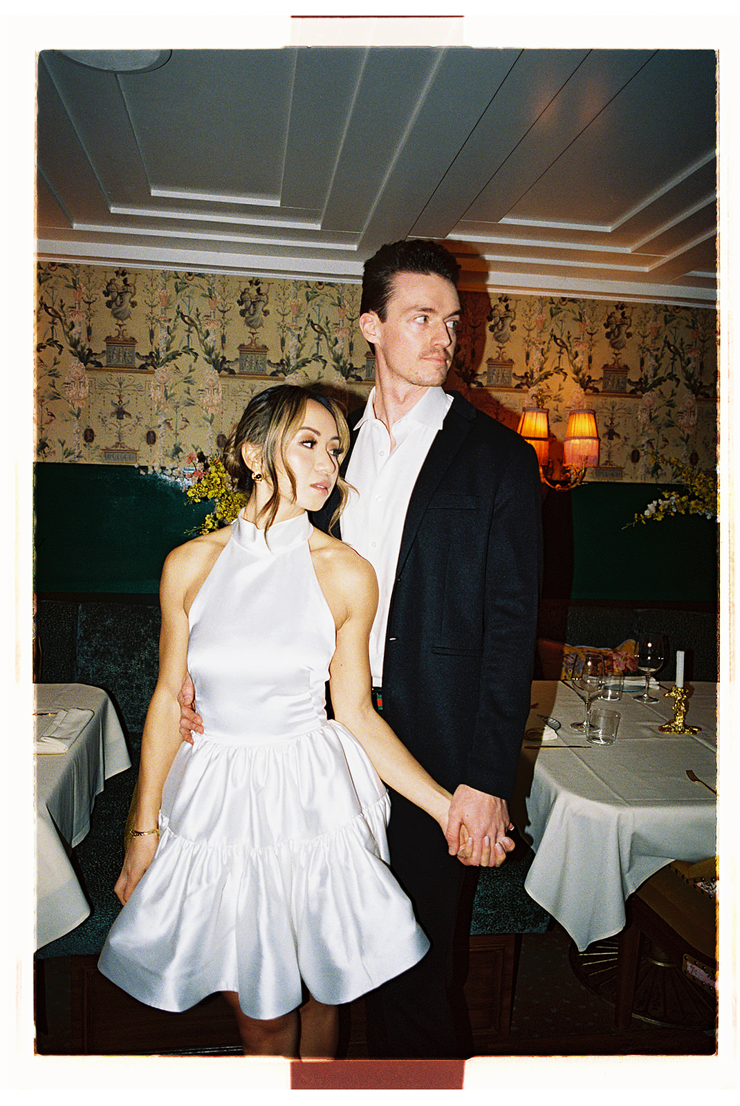 A couple in formal attire shares a dance in a restaurant with white tablecloths and dim lighting.