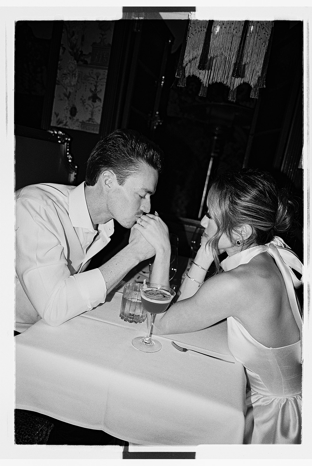 A couple in formal attire shares a dance in a restaurant with white tablecloths and dim lighting.