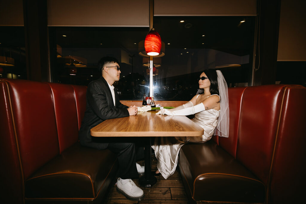 Two people sitting across from each other in a restaurant booth, wearing formal attire and sunglasses, under a red hanging light.