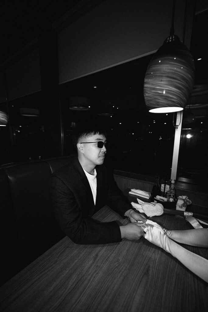 Two people sitting across from each other in a restaurant booth, wearing formal attire and sunglasses, under a red hanging light.