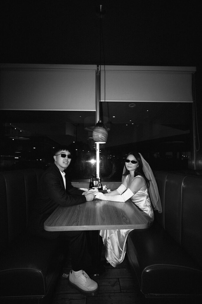 Two people sitting across from each other in a restaurant booth, wearing formal attire and sunglasses, under a red hanging light.