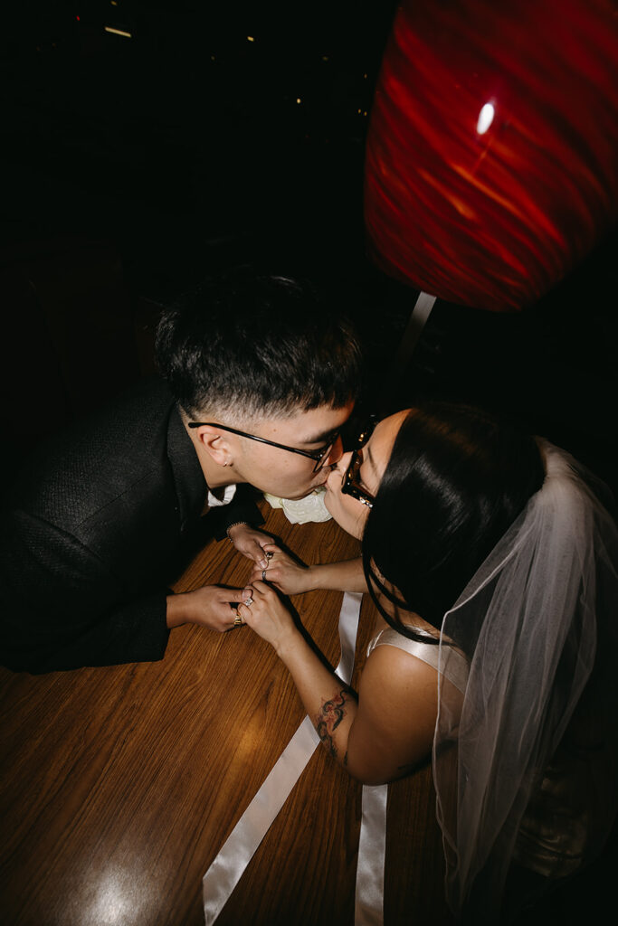 Two people sitting across from each other in a restaurant booth, wearing formal attire and sunglasses, under a red hanging light.