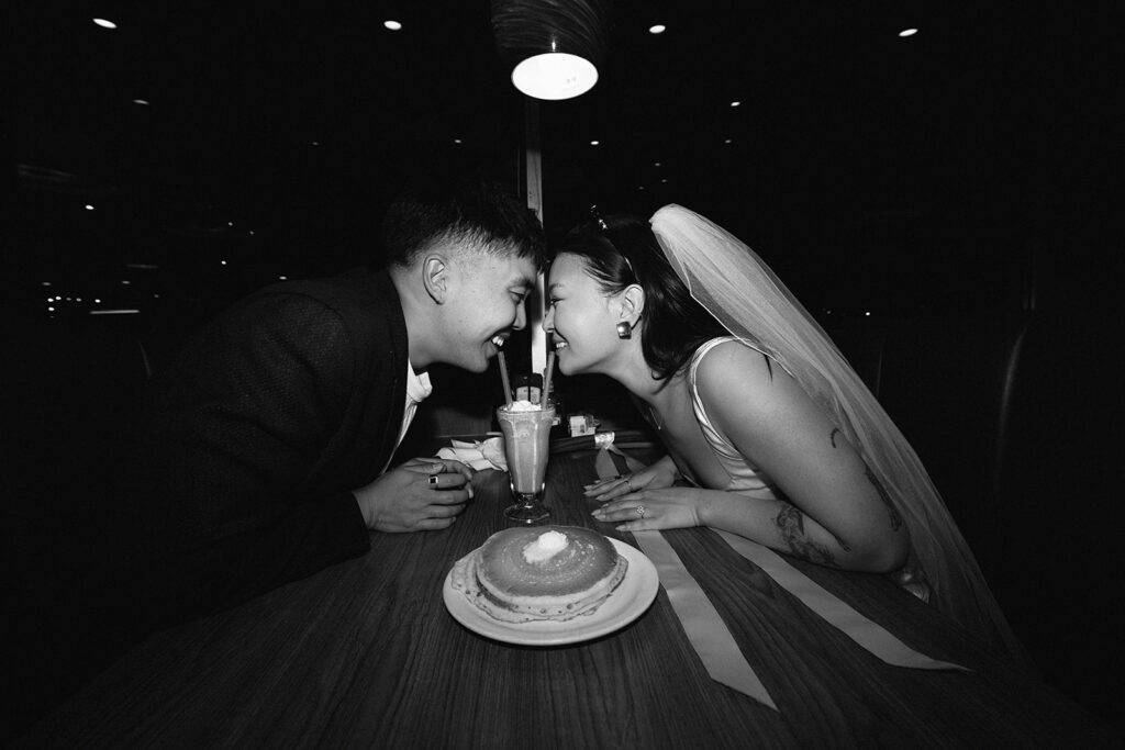 Two people sitting across from each other in a restaurant booth, wearing formal attire and sunglasses, under a red hanging light.